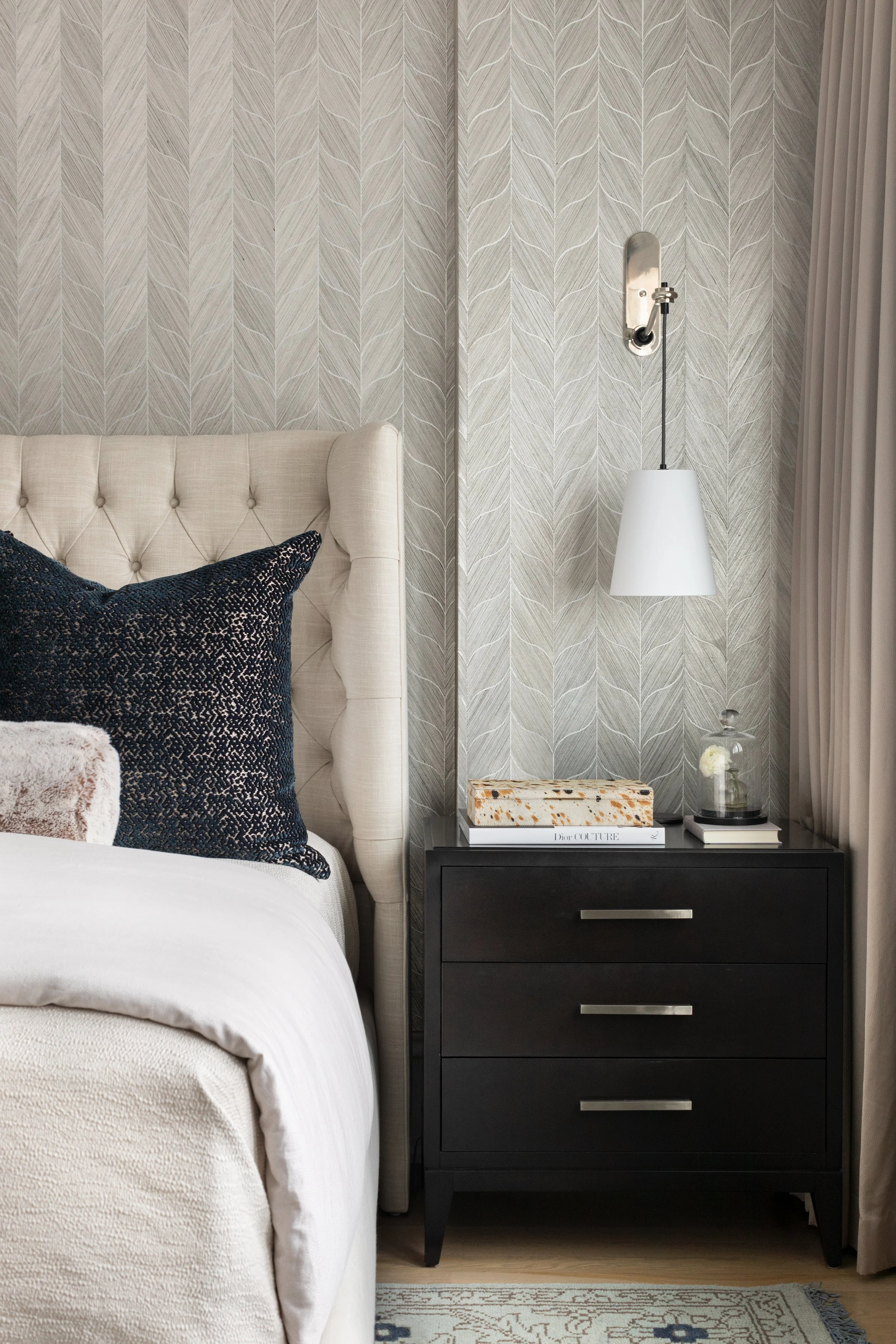 A bedroom corner with a bed, a black nightstand, and a wall-mounted reading light. The bed has a beige tufted headboard and dark blue textured pillows. The nightstand holds a book titled 'Dior Couture,' a decorative box, and a glass dome with a flowe