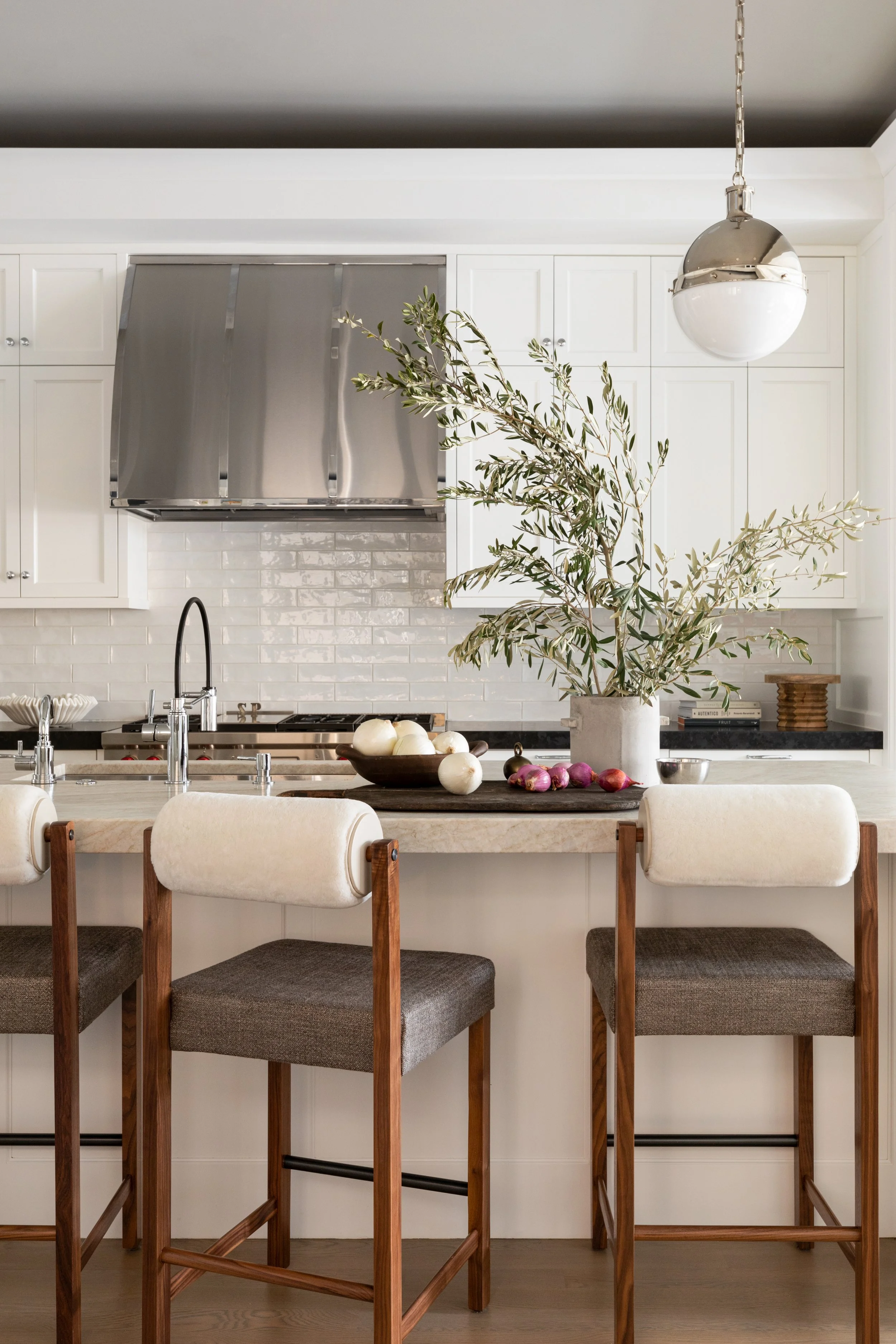 Kitchen with white cabinets, gray backsplash, metal range hood, and a central island with bar stools. A large potted plant with green leaves sits on the island, with vegetables like onions and shallots on a tray, and a bowl of garlic on the counter.