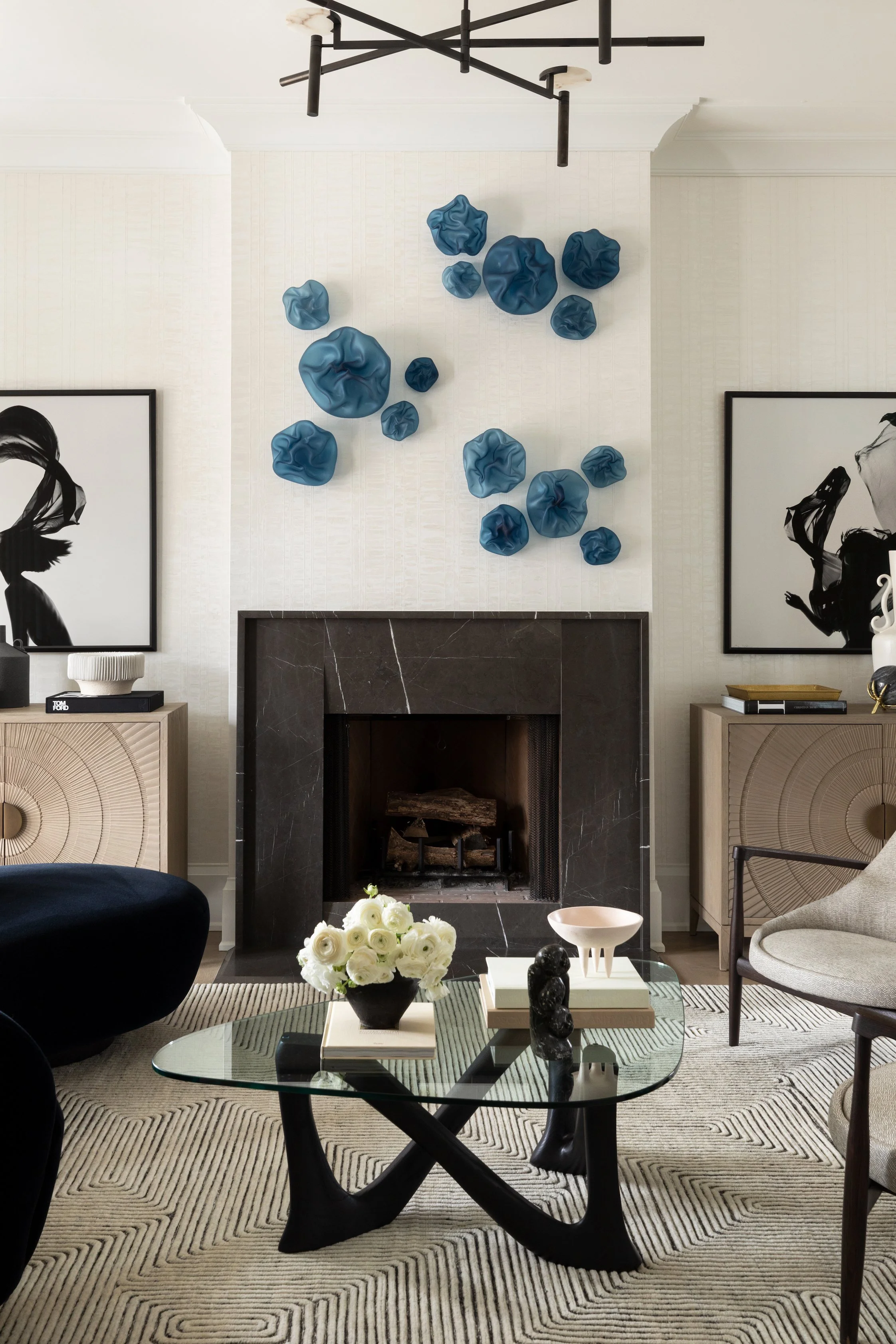 Modern living room with black marble fireplace, white walls, black and white artwork, glass coffee table with a black floral sculpture and white flowers, and a mix of beige and black furniture, including a black velvet chair and a beige armchair. Blu