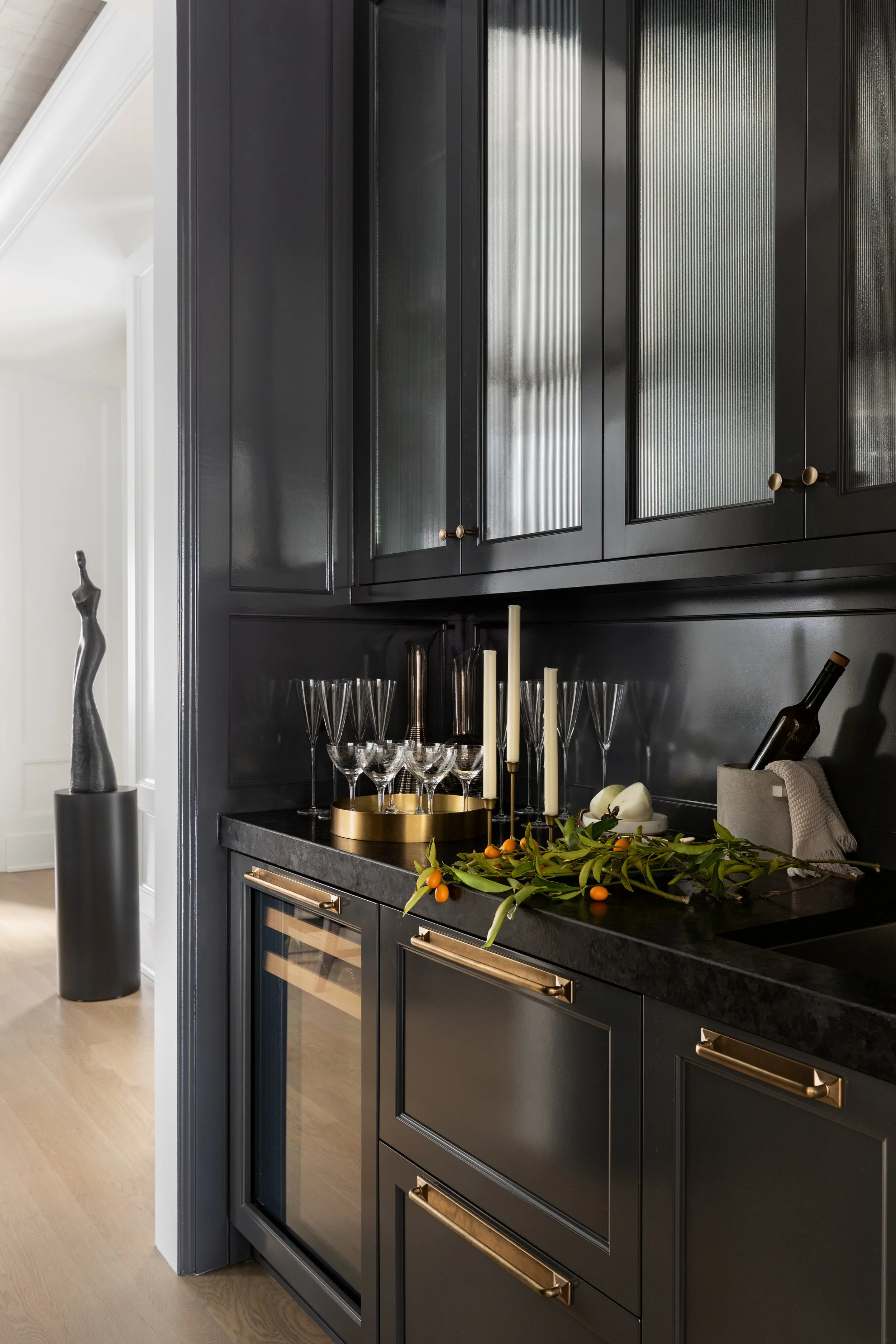 A dark-colored kitchen with black cabinets, gold handles, and black countertop. Decor includes a tray with glasses, tall candles, and a bunch of orange berries with green leaves. A tall black sculpture is on the left, and a wine bottle is in a contai