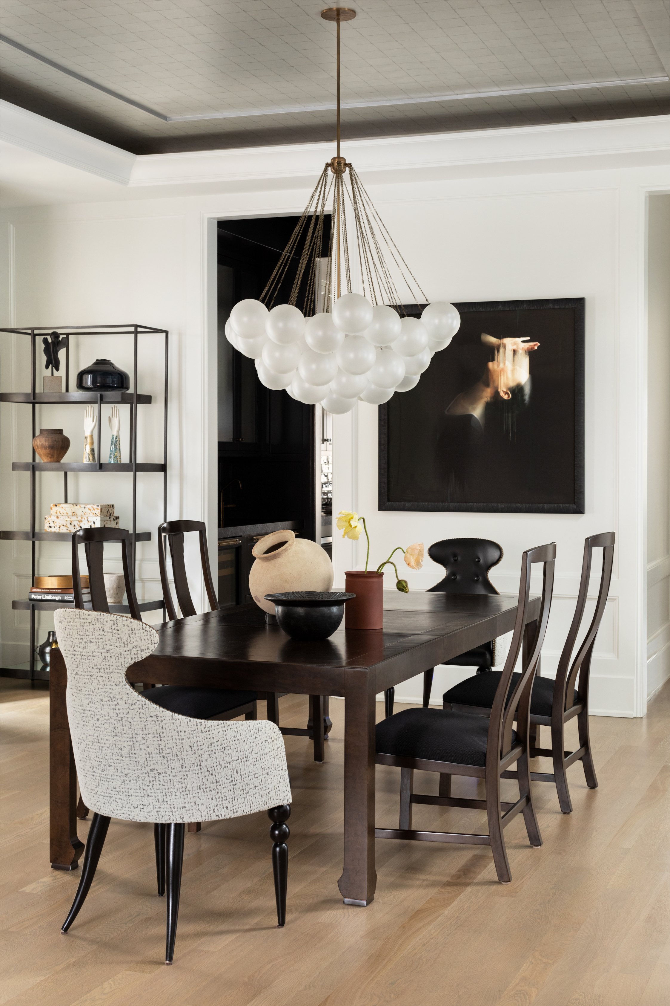A dining room with a dark wooden table, white and black chairs, decorative vases, and a large black-and-white portrait on the wall. A chandelier made of white globes hangs above the table, and a shelving unit displays various decorative items.
