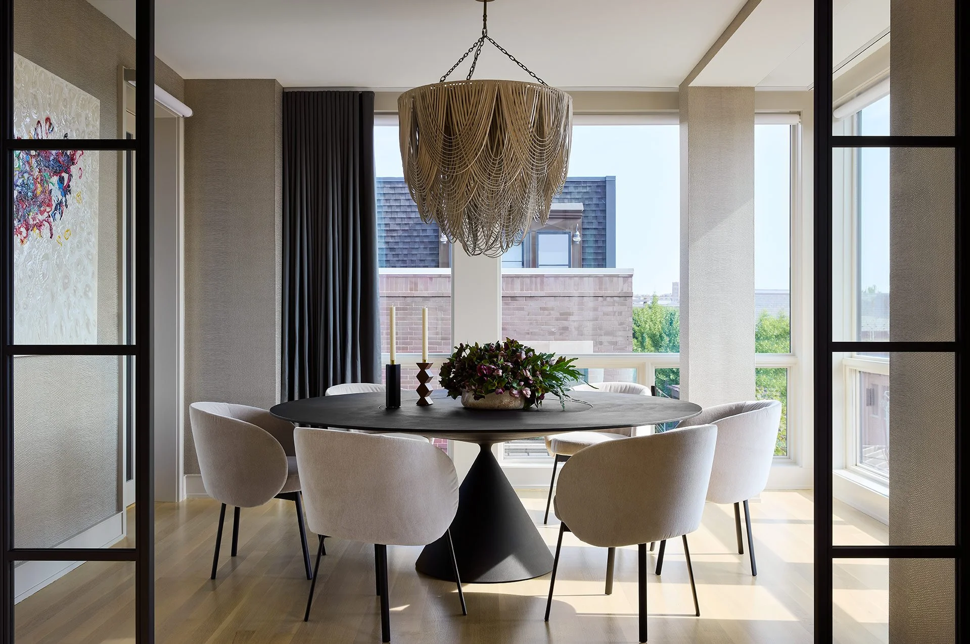 View through a black-framed glass door into a dining room with a round table, six white upholstered chairs, a wooden chandelier, a floral centerpiece, lit candles, and large windows showing a balcony and buildings outside.