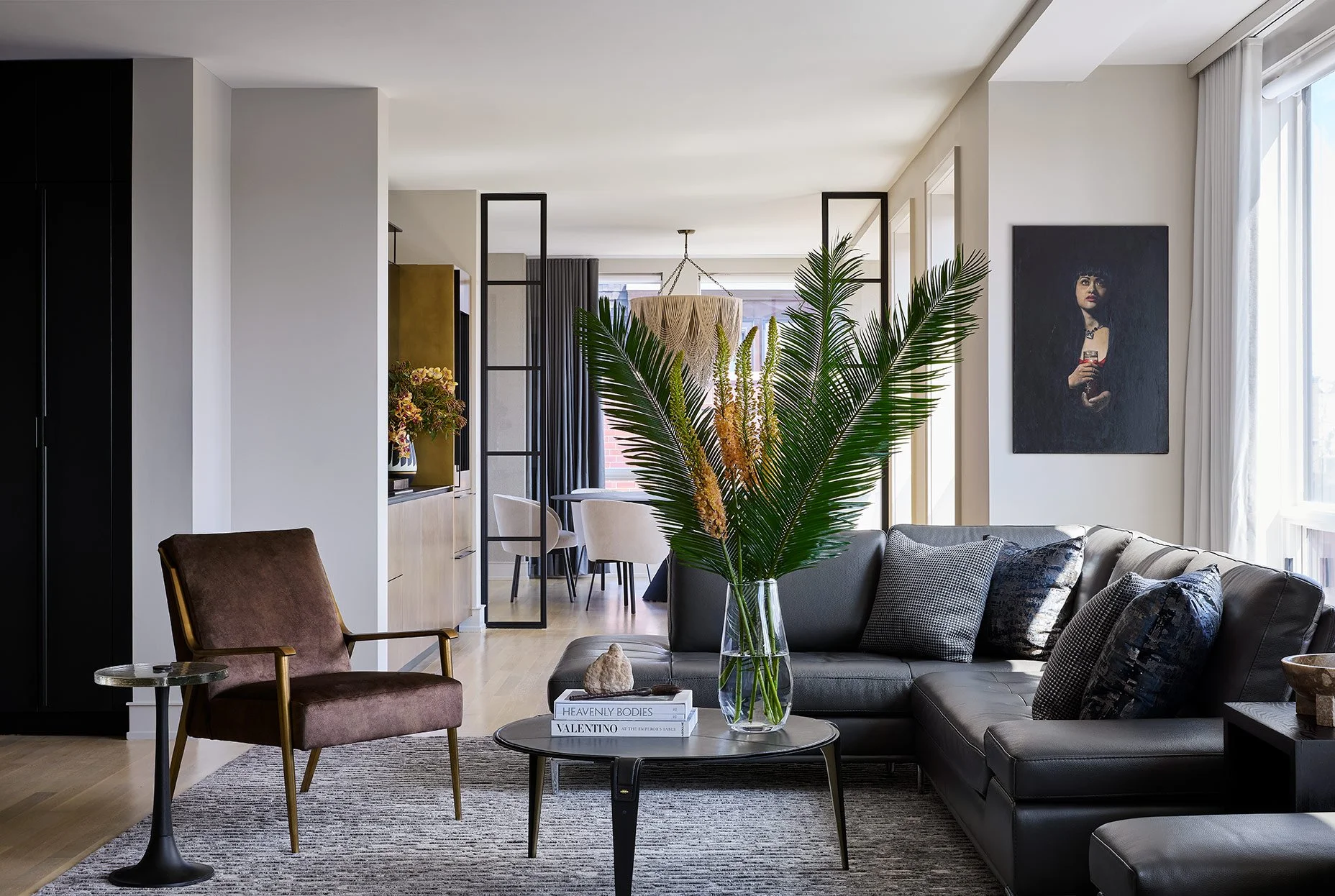Modern living room with black leather sofa, purple armchair, glass coffee table with books, large plant, and artwork on wall, with dining area and window in background.