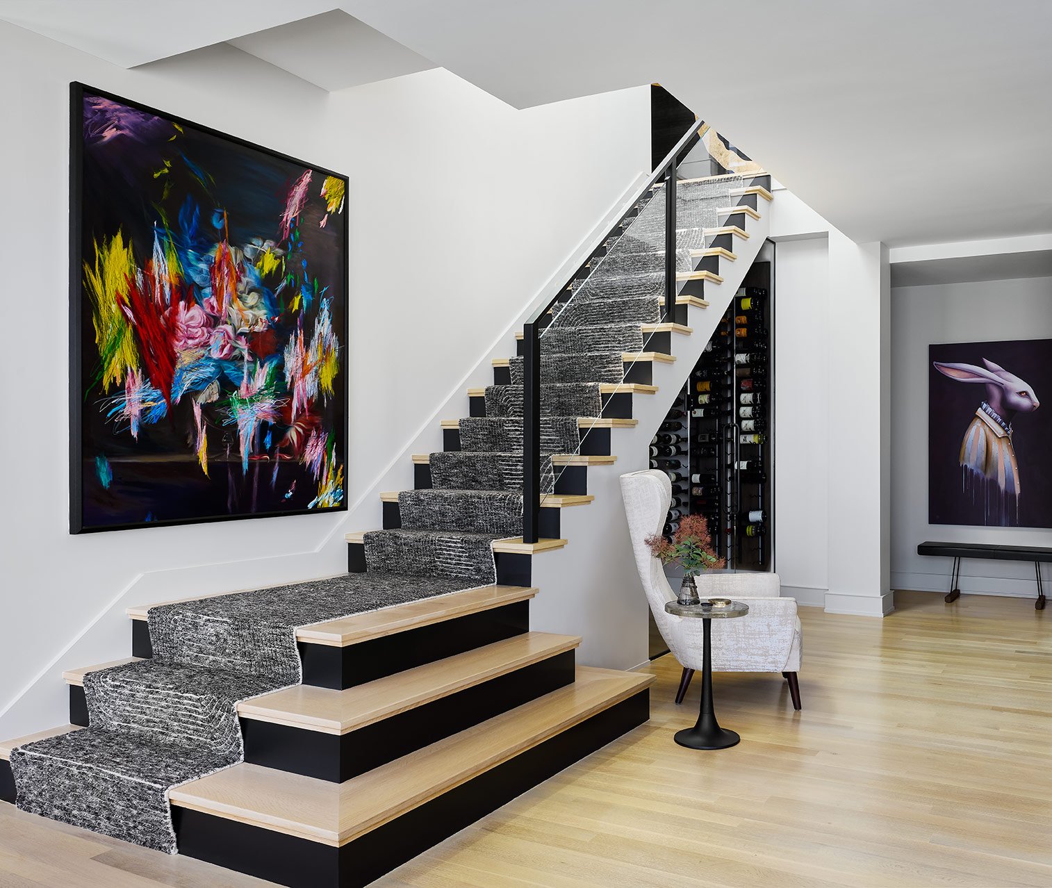 Modern staircase with black and wooden steps and a gray runner rug in a stylish living space. Artworks on the walls, a white high-back chair with a small table and vase with flowers beside it, and a built-in wine rack.