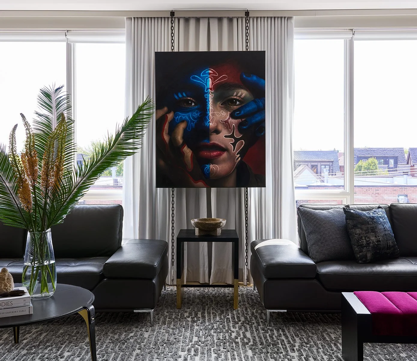 Living room with black leather sofa, decorative pillows, large window with white curtains, abstract portrait painting of a woman with face painted, potted plant on table, and purple ottoman.