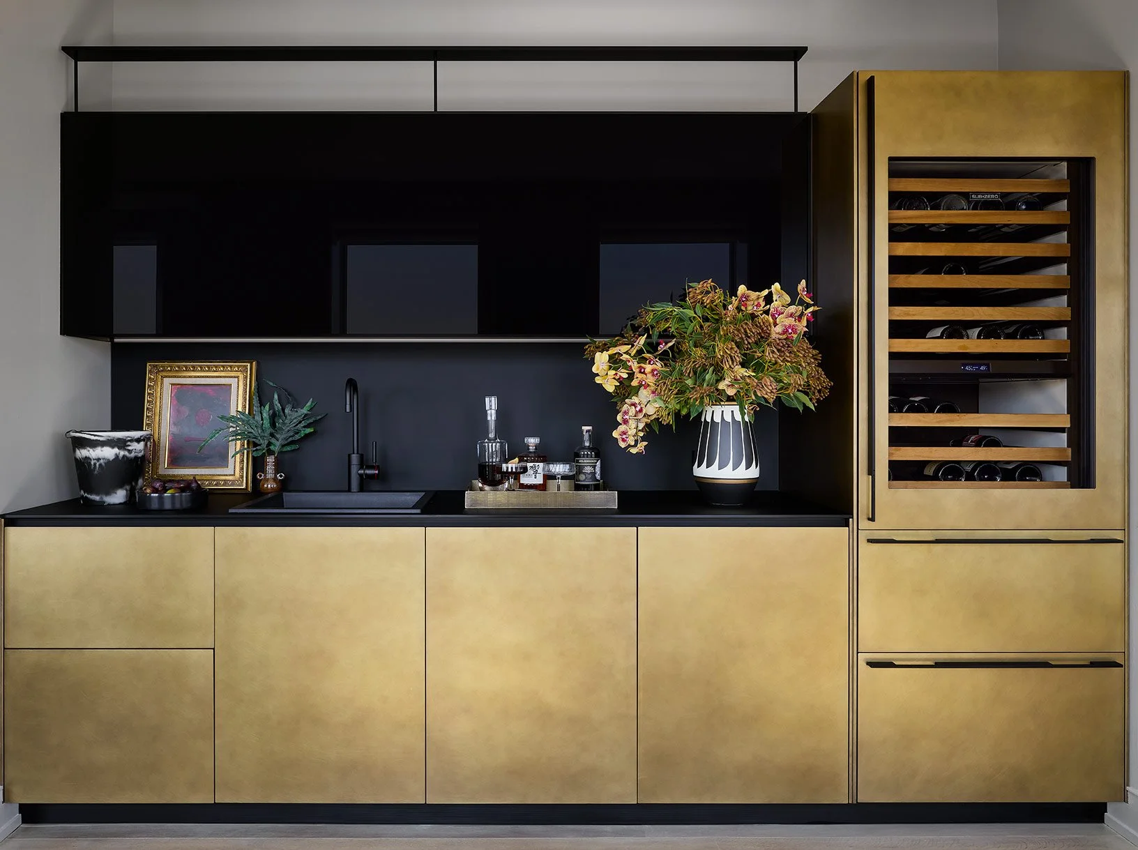 Modern kitchen with black upper cabinets, yellow lower cabinets, a black countertop, a black sink with a black faucet, decorative jars, a framed artwork, a potted plant, and a vase with flowers. A built-in wine cooler is on the right with wooden shel