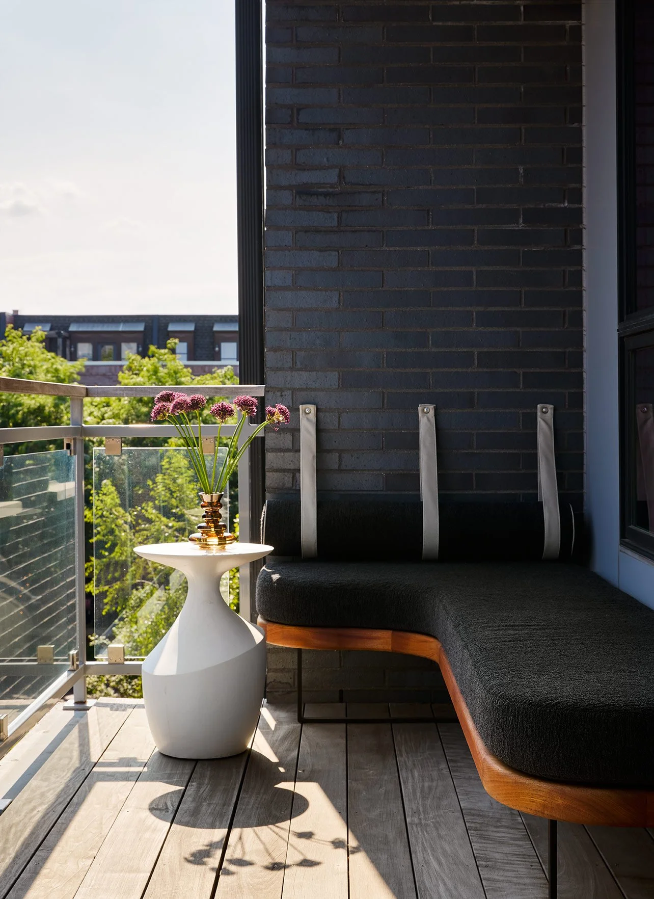 A balcony with wooden flooring, a black brick wall, a modern white side table with a golden vase filled with purple flowers, a dark gray cushioned seating area, and a view of green trees and building rooftops in the background, during daytime.