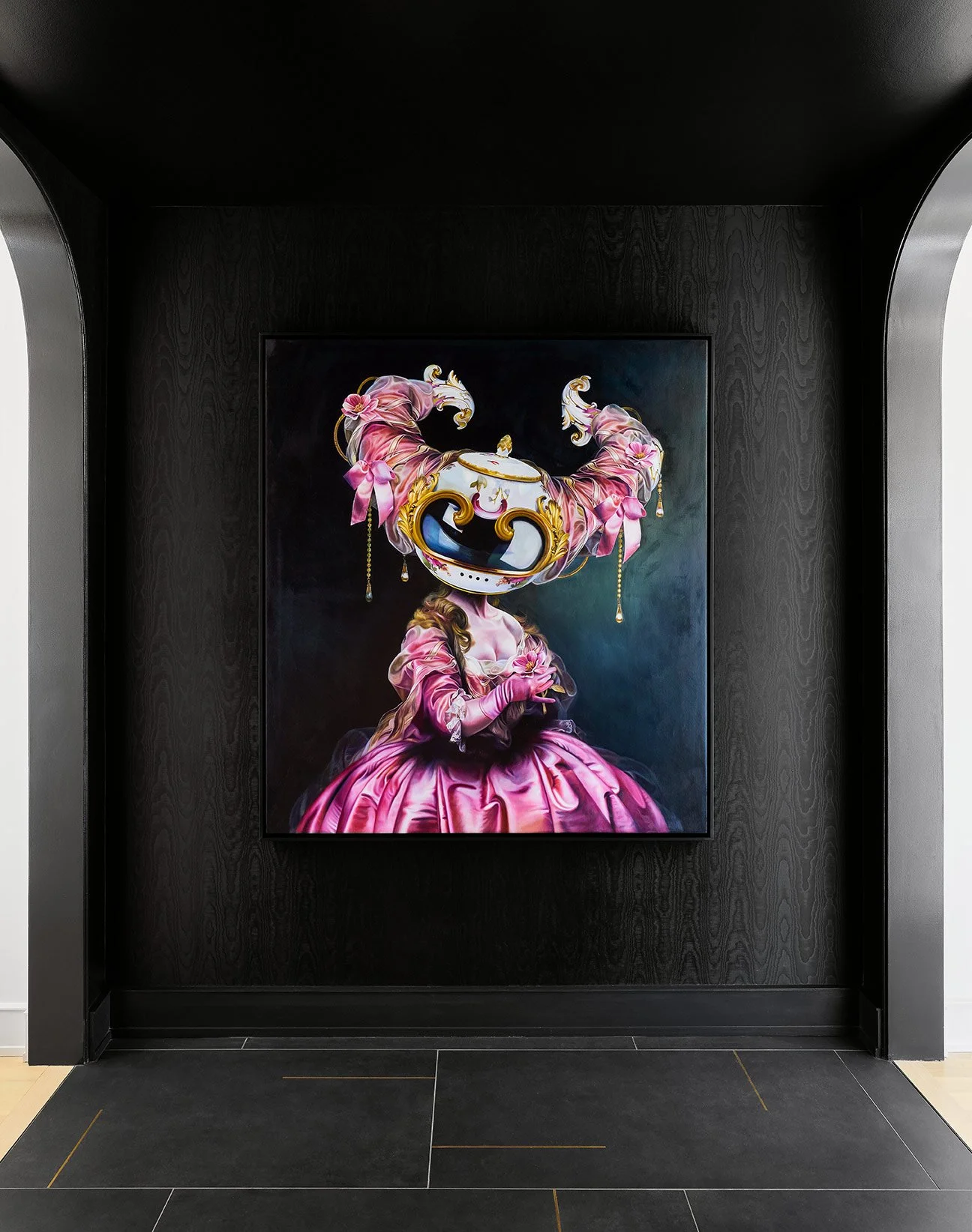 A contemporary art portrait of a woman wearing a pink Victorian-style dress and gloves, with her face replaced by a large ornate mask with decorative ribbons and jewelry, displayed on a dark wall.