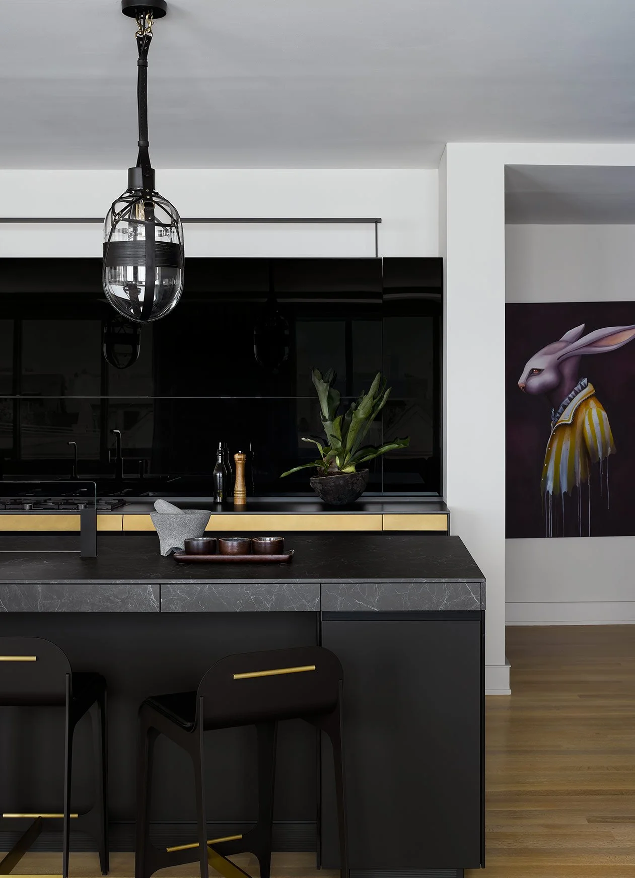 Modern kitchen with black cabinets, a large plant on the countertop, a hanging black and glass light fixture, and a colorful rabbit painting on the wall.