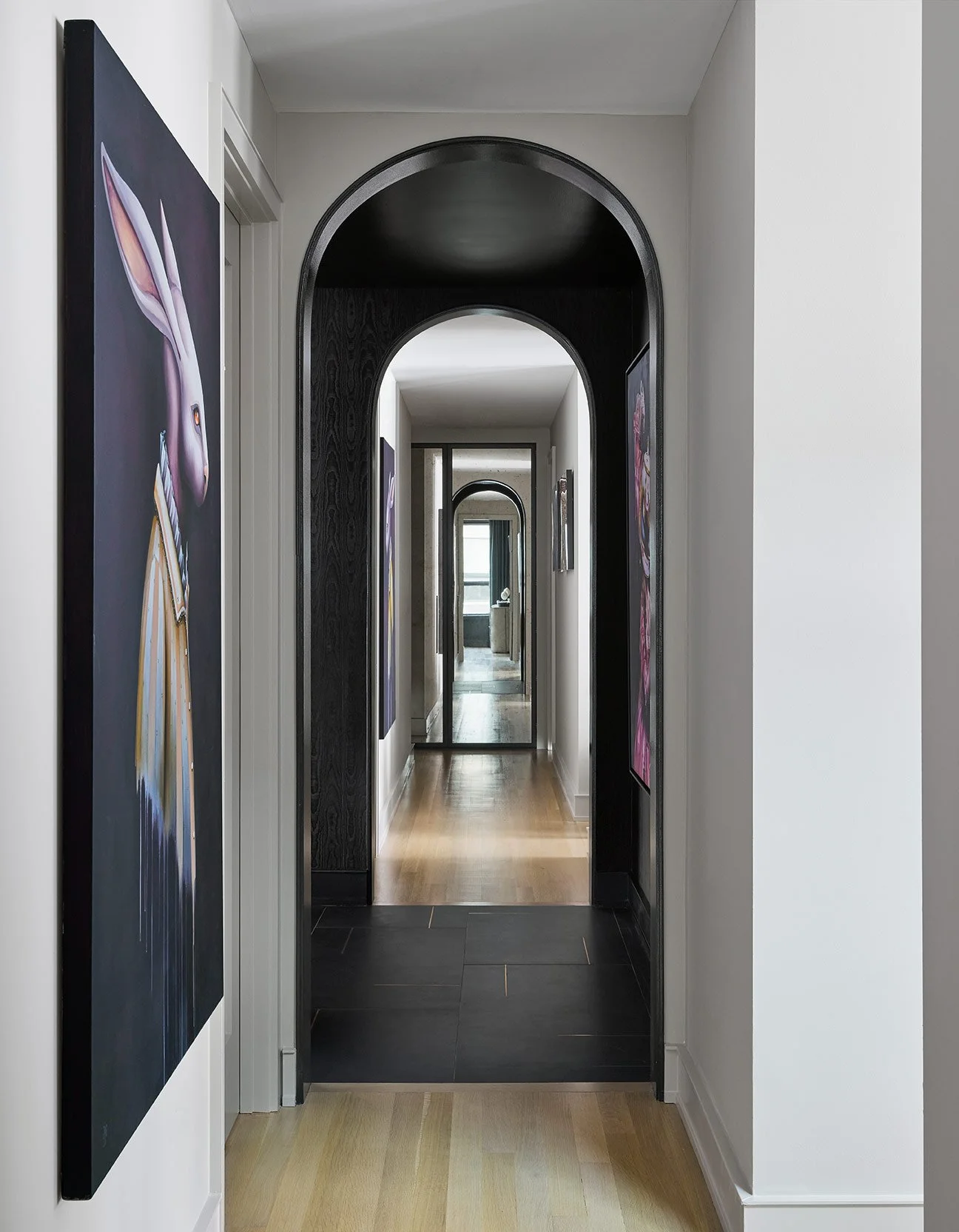 Hallway with artistic paintings on both sides, black and white archways, and a mirror at the end reflecting a window.