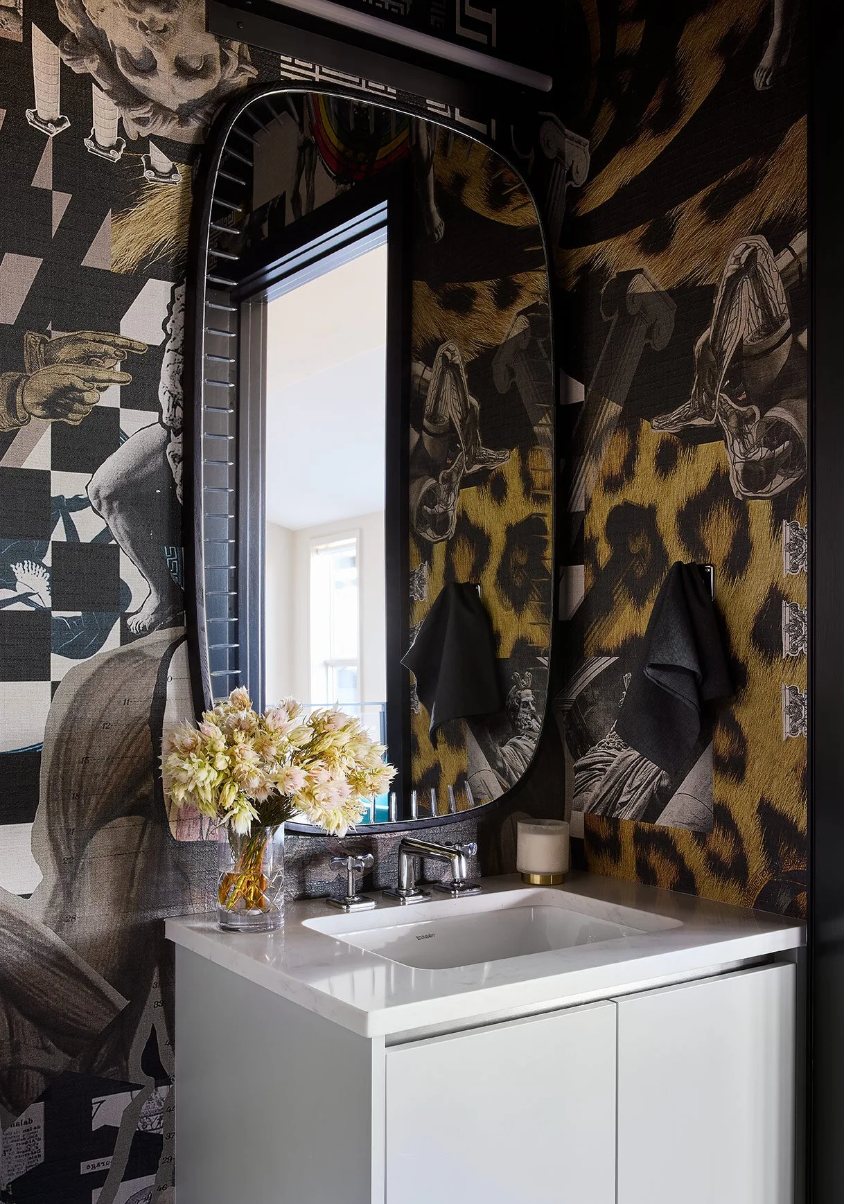 Bathroom vanity with a white sink, two black towels hanging on mirrored cabinet doors, a vase of pink and cream flowers, a soap dispenser, and a window reflected in the mirror, all against a wall decorated with black, gold, and grayscale mural artwor