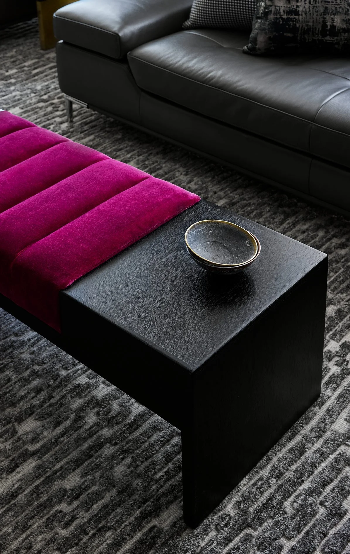 Close-up of a modern living room with a black wooden coffee table holding a black bowl, a gray leather sofa, and a pink velvet bench, all on a patterned gray carpet.