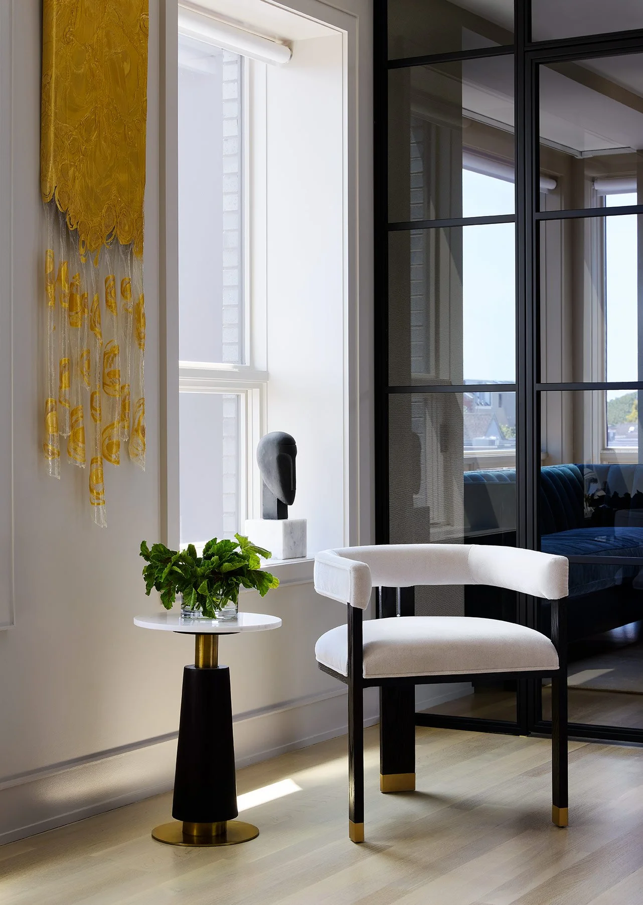A modern interior with a white chair, a side table with a green plant, and decorative art on a window ledge, separated by a black-framed glass wall.