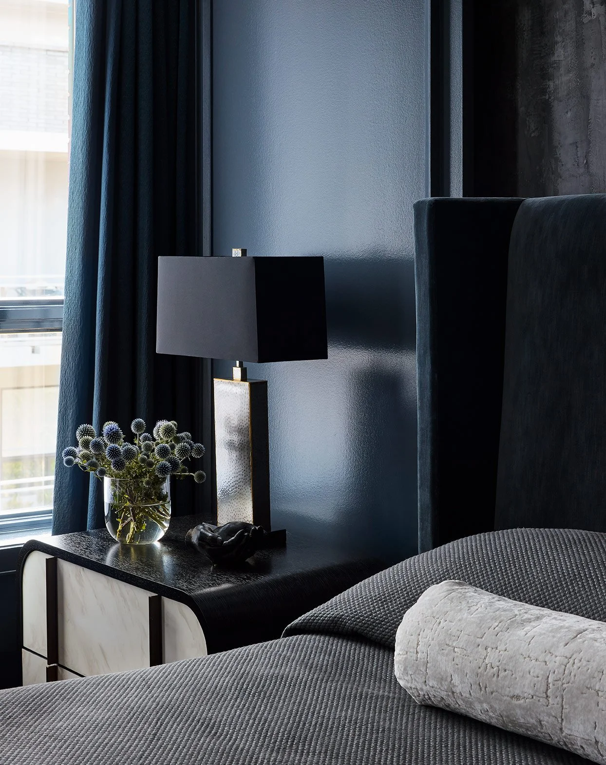 A modern bedroom corner featuring a bedside table with a black and gold lamp, a vase of purple thistle flowers, and a black decorative object. A bed with a gray quilt and a rolled light-colored pillow is in the foreground. The paneled wall has blue a