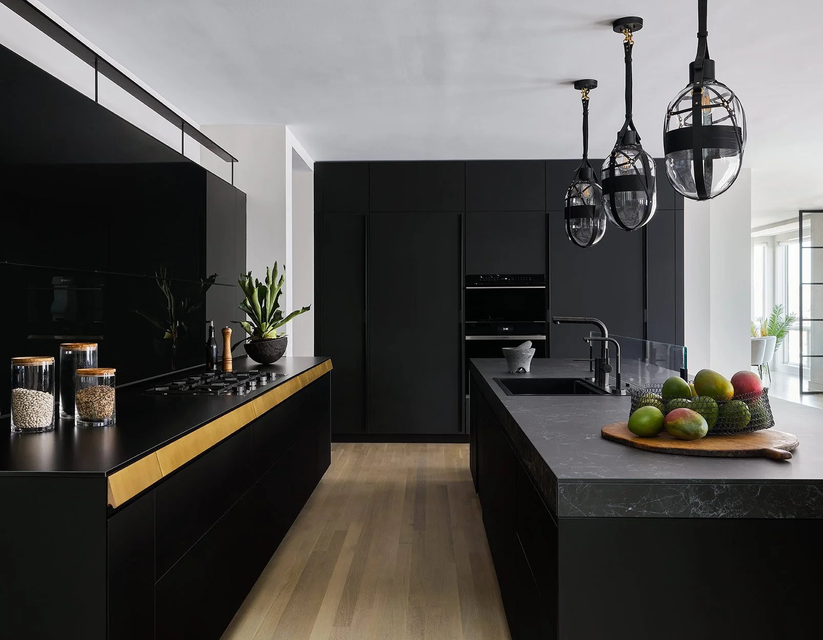 Modern black kitchen with gold accents, hanging pendant lights, green mangoes on a black marble island, and a potted plant on the counter.