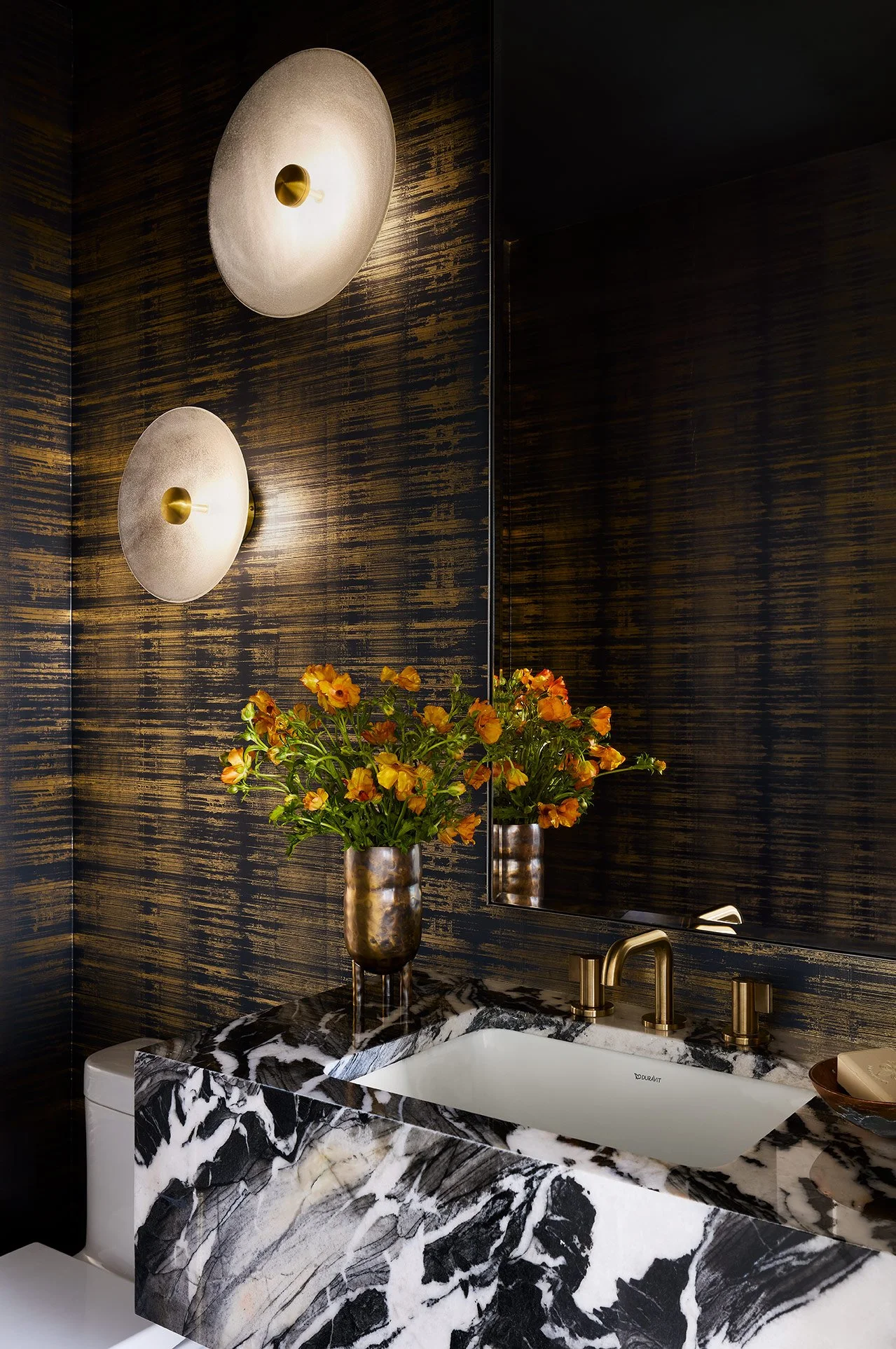 A bathroom vanity with a black and white marble countertop, a gold faucet, a large mirror, two wall sconces, and a vase with orange flowers on a dark textured wall background.