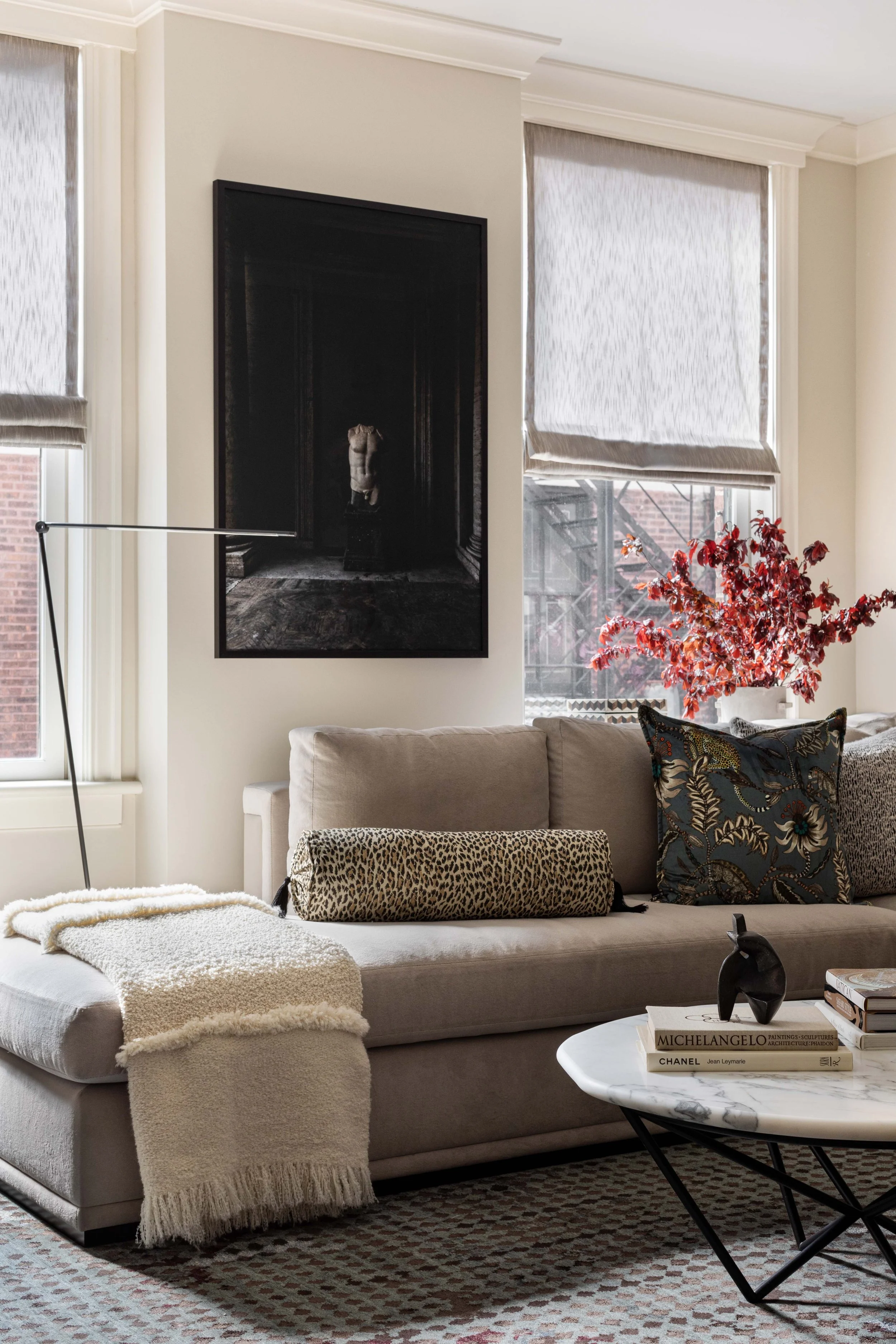 Living Room with beige sectional sofa, patterned pillows, a white marble coffee table with books and decorative objects, large windows with shades, a framed artwork with a torso sculpture, a leopard-print pillow, and a red-leaved plant, in Chicago.