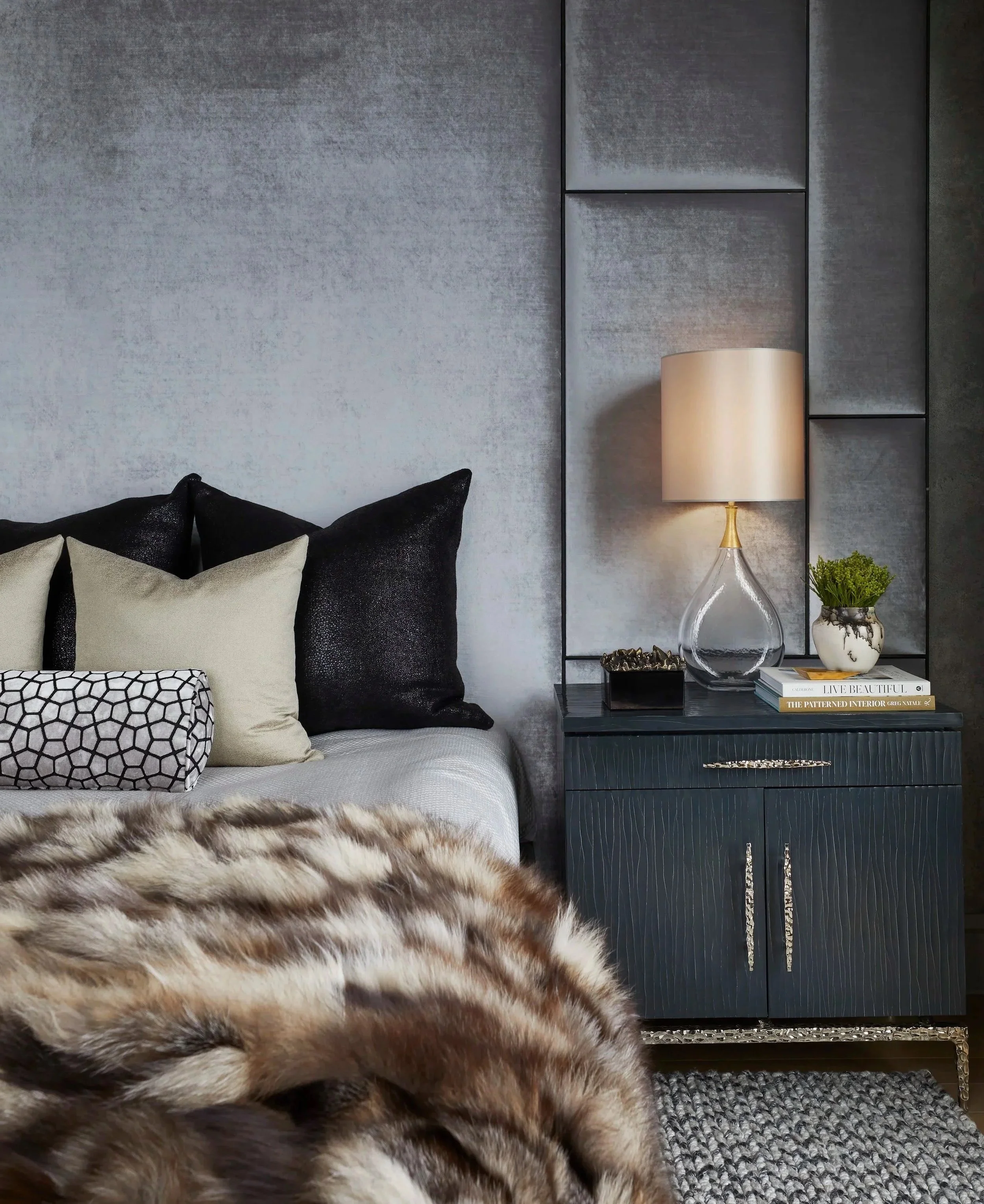 A primary bedroom with a full wall velvet headboard, faux fur throw blanket, patterned cushions, a table lamp with a cream shade, a small plant in a white pot, and a glossy black dresser with a decorative box and books. All designed by Donna Mondi.