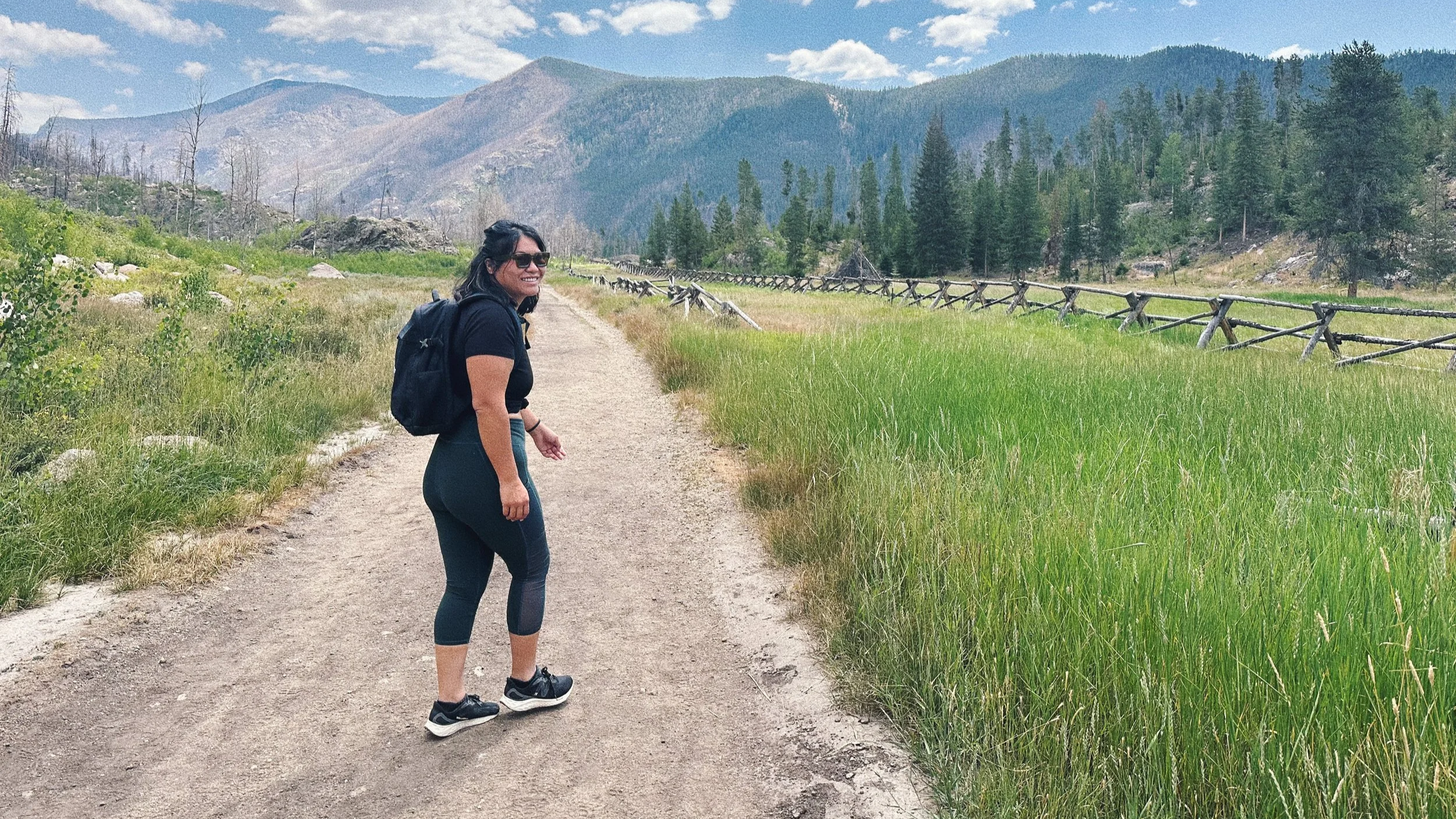 A woman with dark hair, wearing sunglasses, a black t-shirt, and leggings, is smiling and walking along a dirt trail in a lush green meadow. She has a backpack and is surrounded by trees, mountains, and a partly cloudy sky.