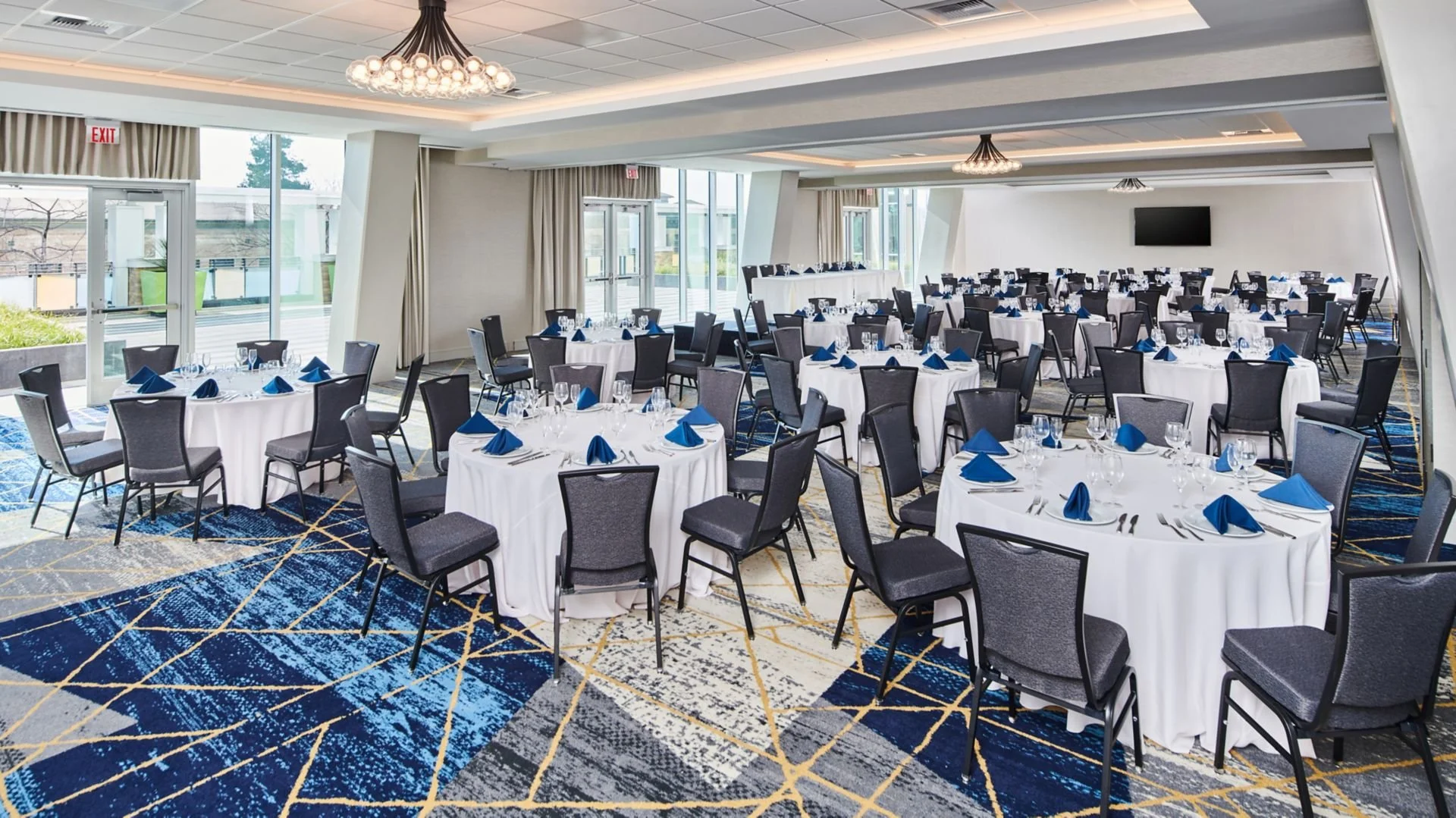 A banquet hall with round tables set for an event, with white tablecloths, blue napkins, glassware, and silverware, and large windows and glass doors providing natural light.