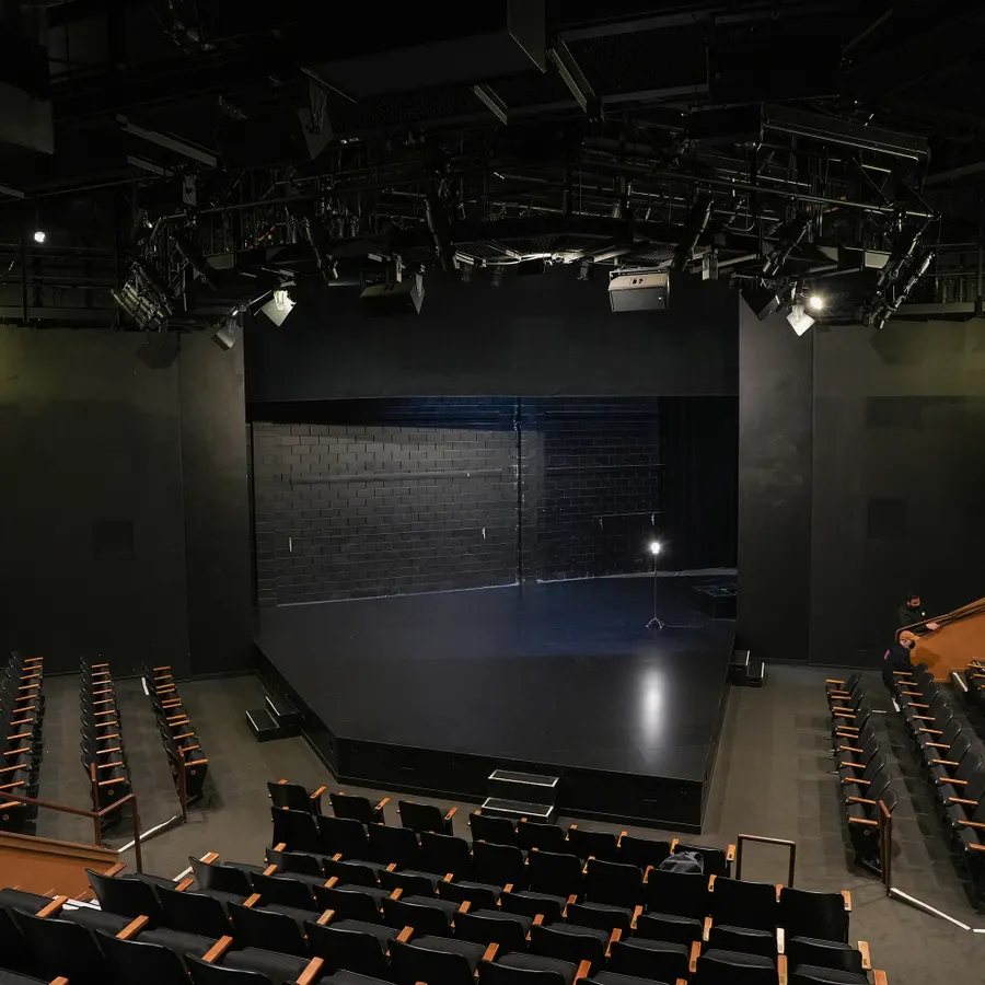 Empty theater stage with black walls and curtains, surrounded by rows of black seats with wooden armrests, illuminated by stage lighting equipment overhead.