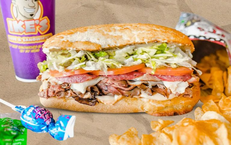 Sub sandwich with lettuce, tomato, bacon, turkey, and roast beef on a toasted bun, with a side of potato chips and a purple drink in the background.