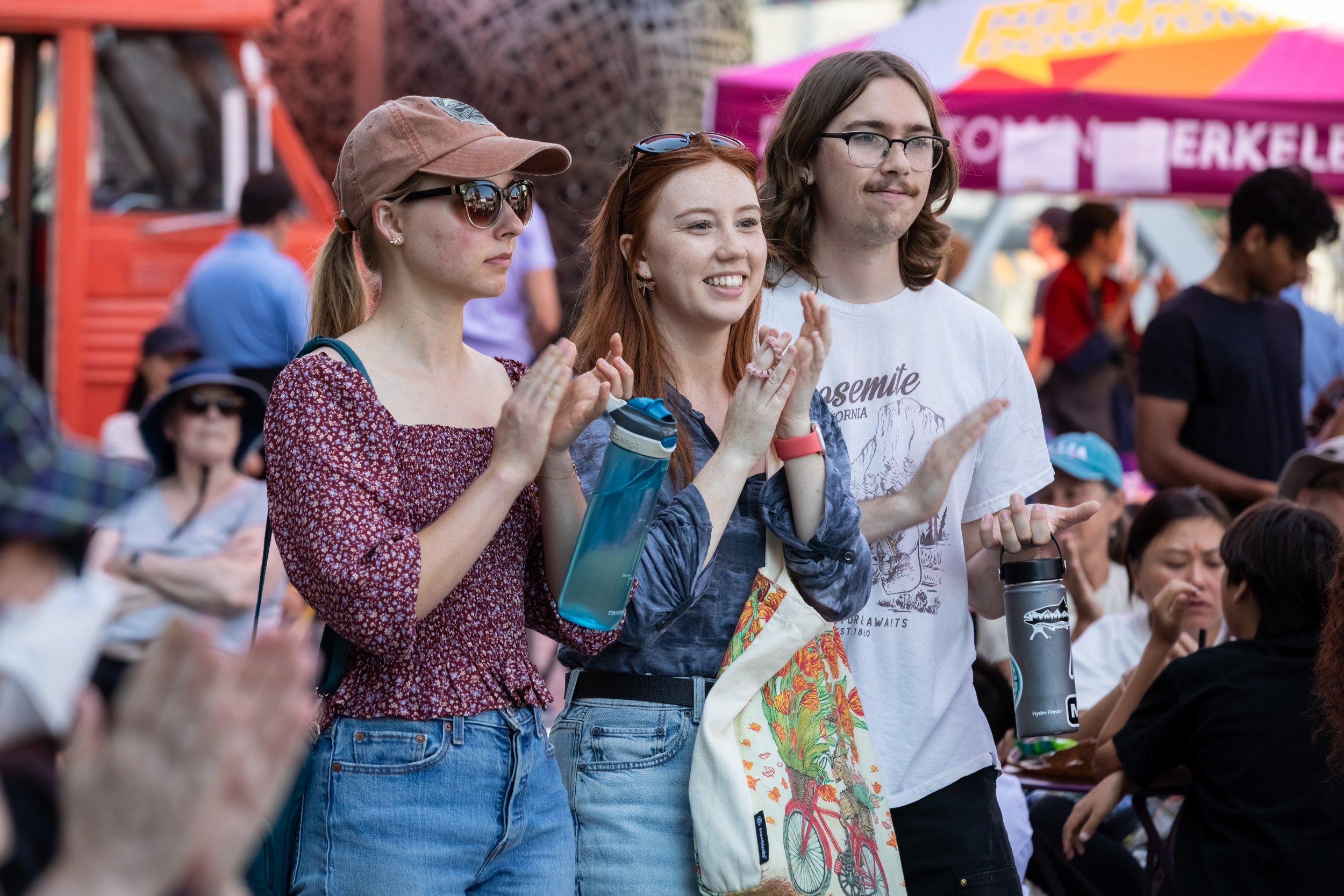 Three young adults are standing in a crowd at an outdoor event. The woman on the left wears sunglasses, a tan cap, and a red dress with small white flowers. She carries a blue water bottle. The woman in the middle has red hair, freckles, and wears earrings and a gray jacket. She is smiling and clapping. The man on the right has long brown hair, glasses, facial hair, and is wearing a white t-shirt with a graphic print. They are surrounded by other people and there is a purple banner in the background.