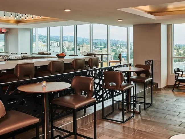 Interior of a modern lounge or restaurant with bar seating, leather chairs, round tables, large windows with city skyline view, and warm lighting.