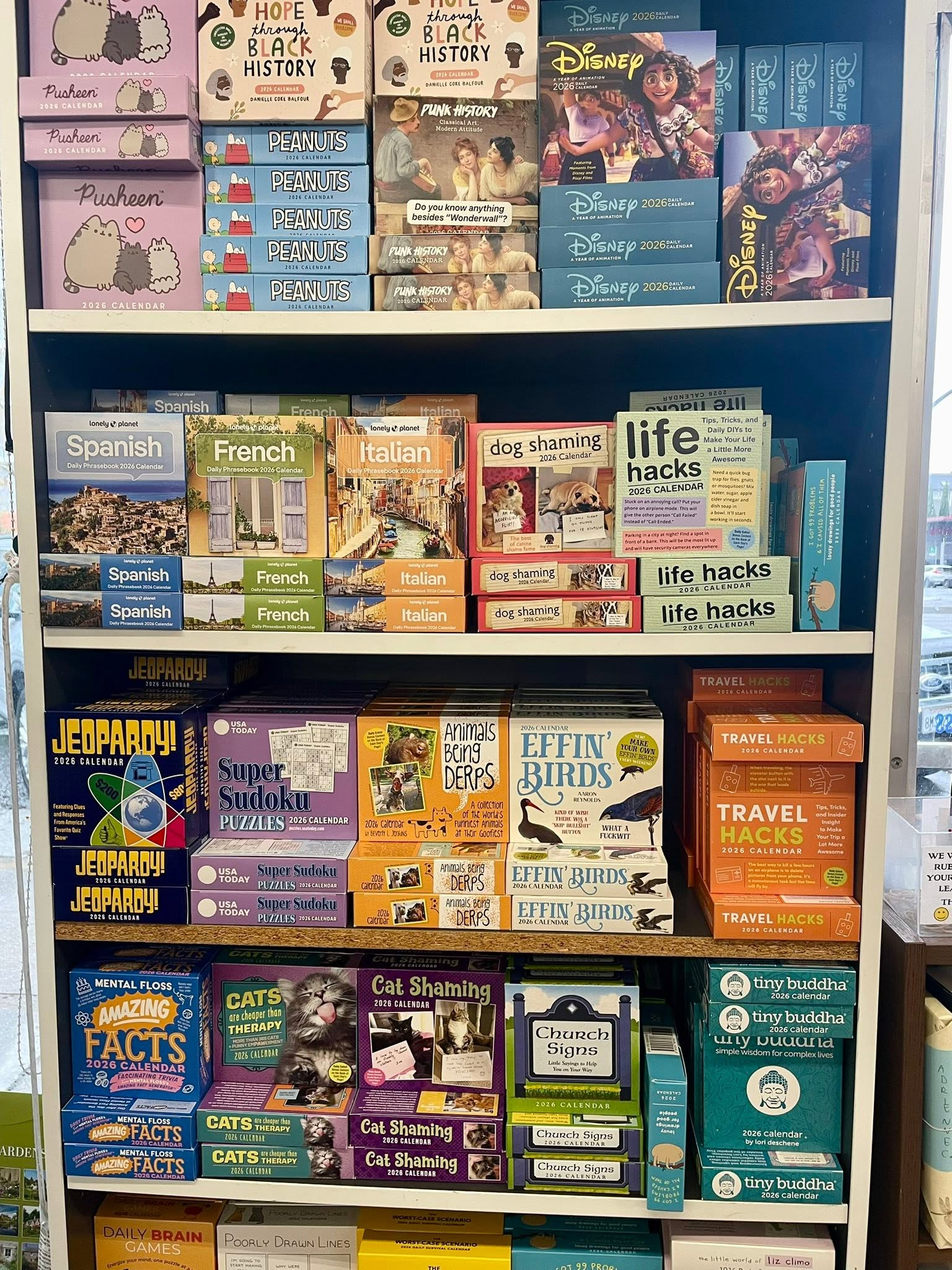 Shelf of 2026 calendars including titles like 'Pusheen', 'Peanuts', 'Disney', 'Spanish', 'French', 'Italian', 'Dog Shaming', 'Life Hacks', 'Jeopardy!', 'Super Sudoku', 'Animals Being Derps', 'Effin' Birds', 'Travel Hacks', 'Cat Shaming', 'Church Signs', 'Tiny Buddha', and 'Mental Floss'.