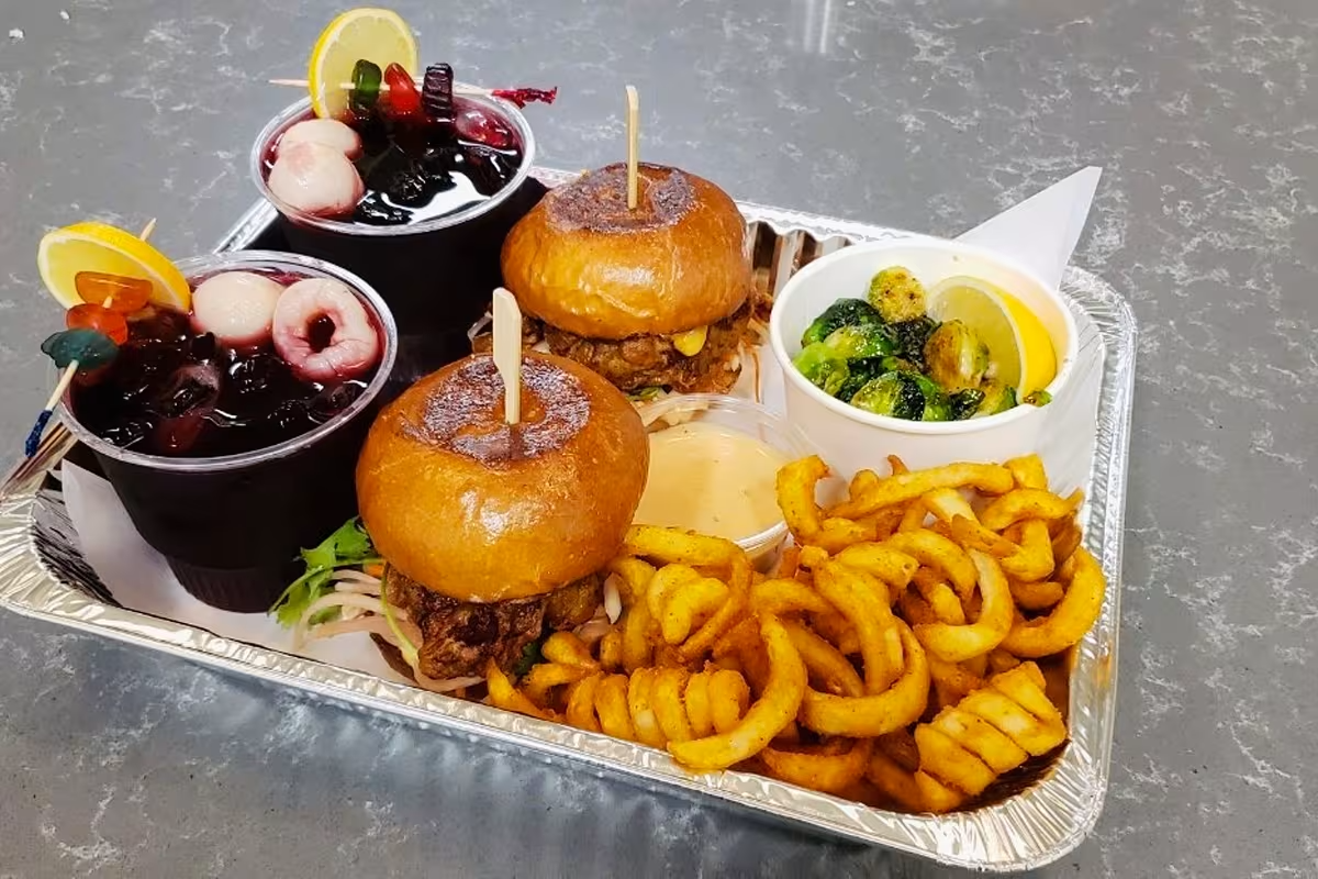 Tray with two mini cheeseburgers with onion and lettuce, curly fries, a cup with Brussels sprouts and lemon wedge, and two cups of fruit cocktails with cherries, grapes, and lemon slices
