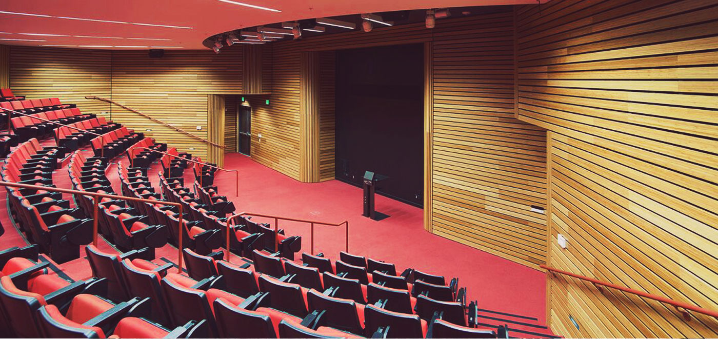 Empty auditorium with red chairs, wooden panel walls, and a large black screen at the front.