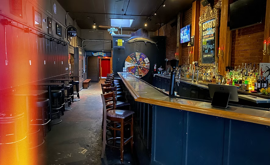 Interior of a bar with a long wooden counter, chairs, bar equipment, a large mounted manta ray, a spinning wheel, and wall-mounted screens, with brick and wood decor.