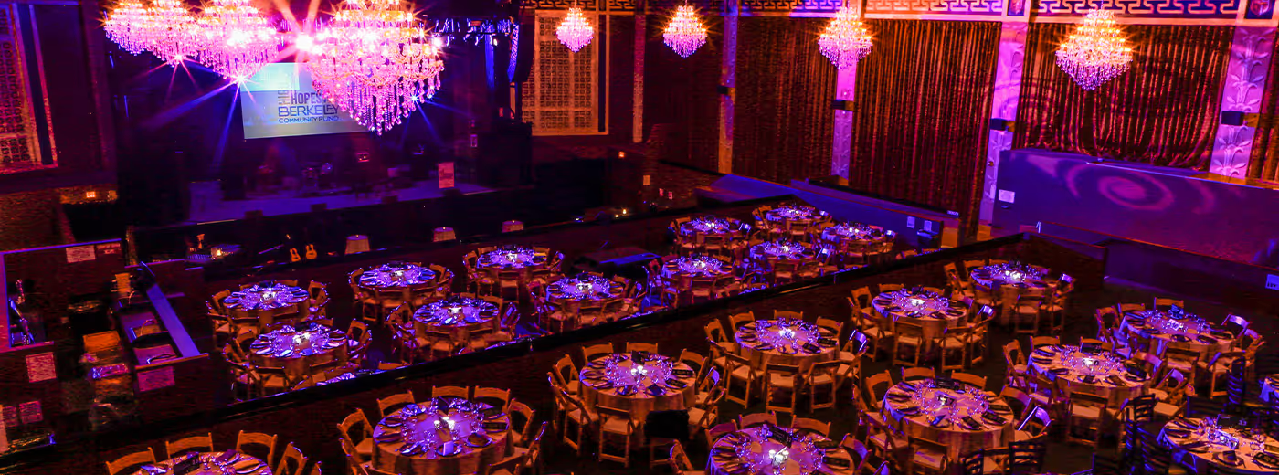 Elegant banquet hall decorated with large chandeliers, purple lighting, round tables with place settings, and a stage with a large digital screen.