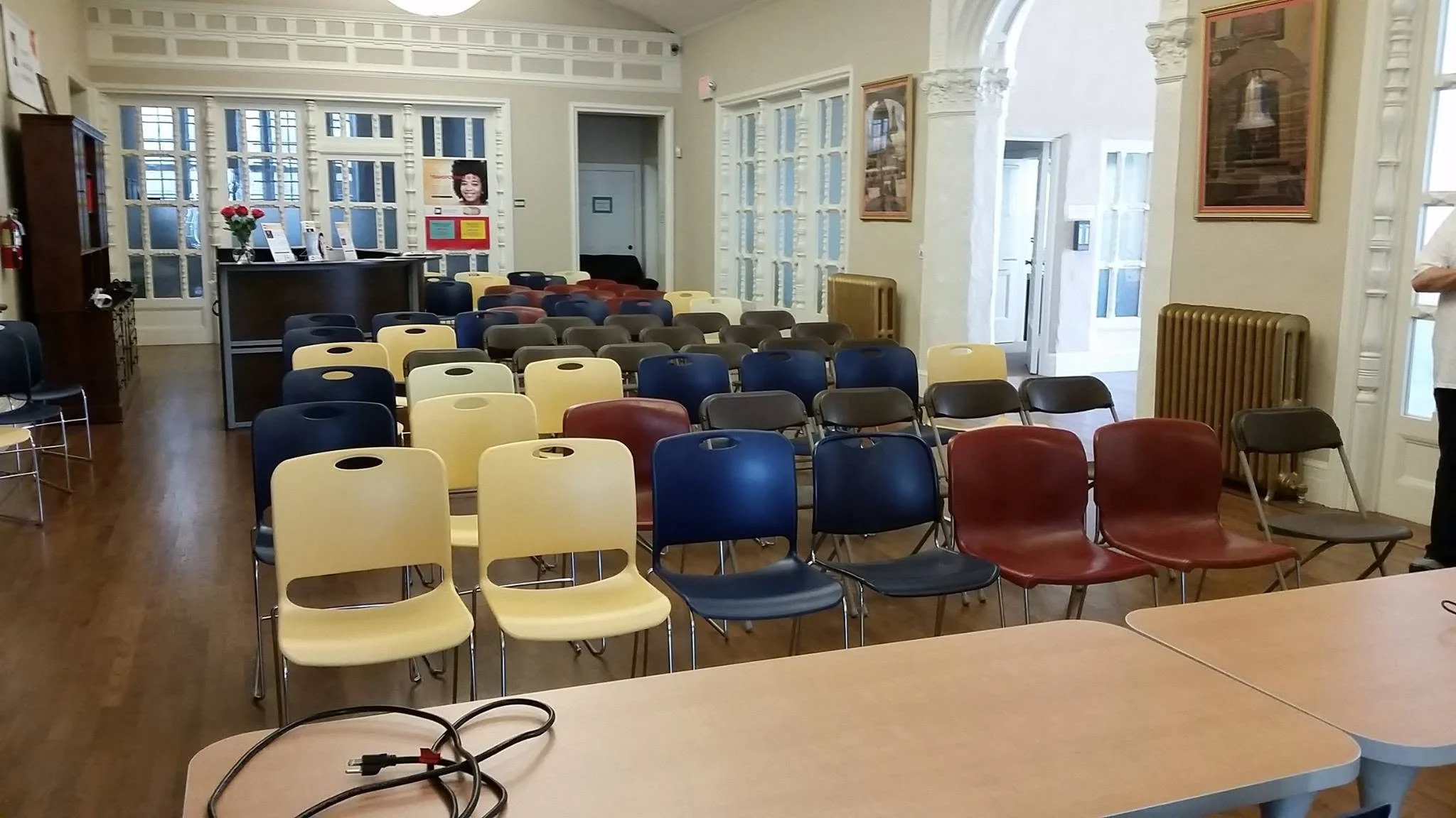 An empty room set up for a presentation or lecture with rows of multicolored chairs, a table with cables, and a front desk area. There are framed pictures and a poster on the walls, and the room has large windows letting in natural light.