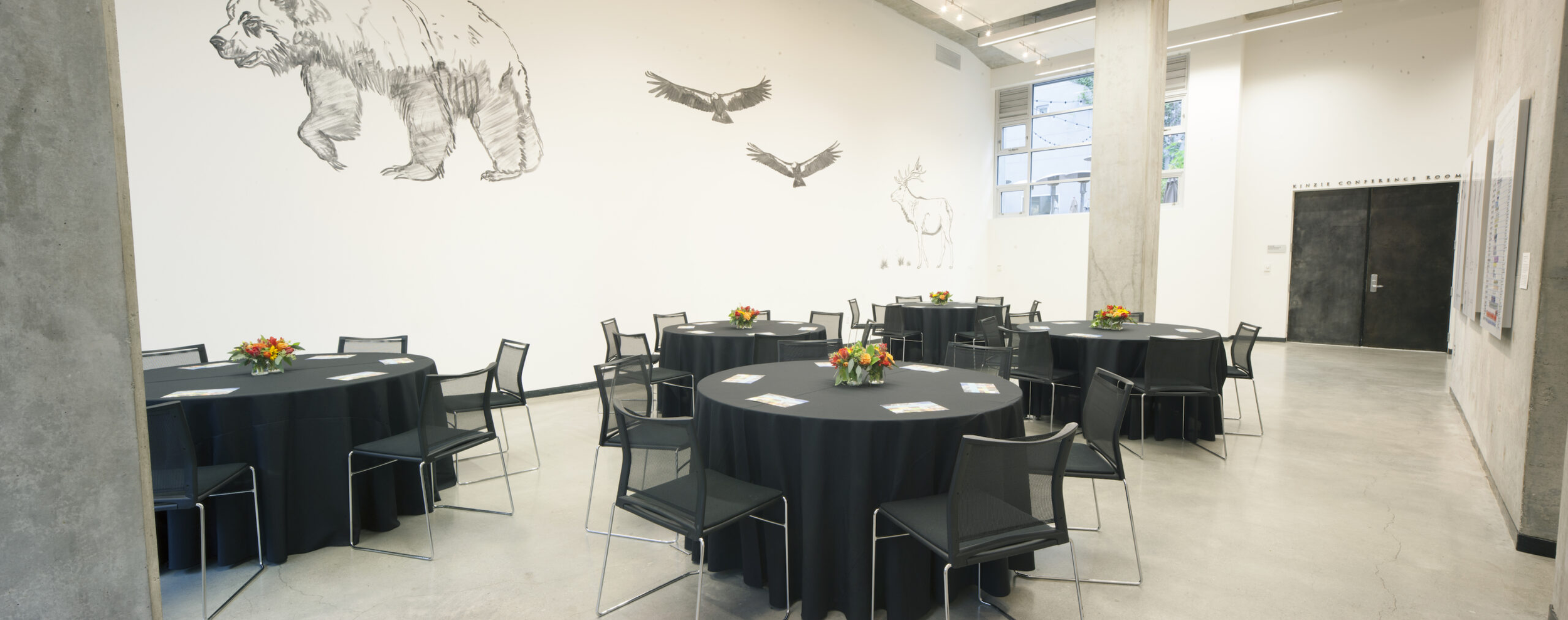 An event space with round tables covered in black tablecloths, each decorated with a small floral centerpiece, scattered with papers and pens, and surrounded by black chairs; the wall features sketches of animals including a bear, birds, and a deer, and the room has large windows letting in natural light.