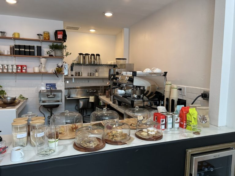Coffee station and baked goods display at a cafe with glass cake covers, coffee machines, cups, and various pantry items.