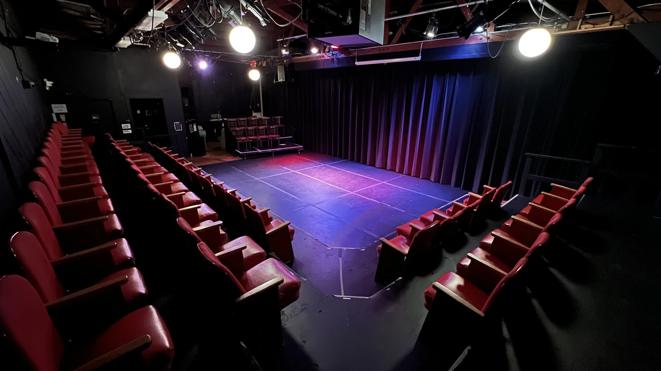 An empty small theater or comedy club with a stage, red chairs, and black curtains.