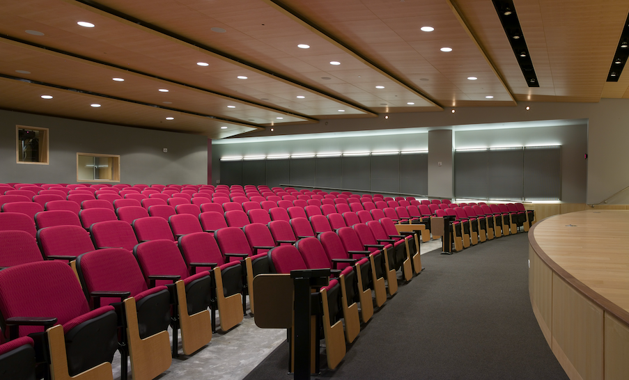 An empty auditorium with rows of red theater seats and a curved wooden stage. The ceiling has recessed lighting and wood paneling.