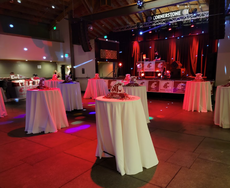An indoor event space decorated with white cocktail tables draped with cloth, each with a red and white centerpiece featuring the Washington State Cougars logo. There is a stage with a DJ setup, red curtains, and lighting. The atmosphere is festive, likely for a party or celebration.