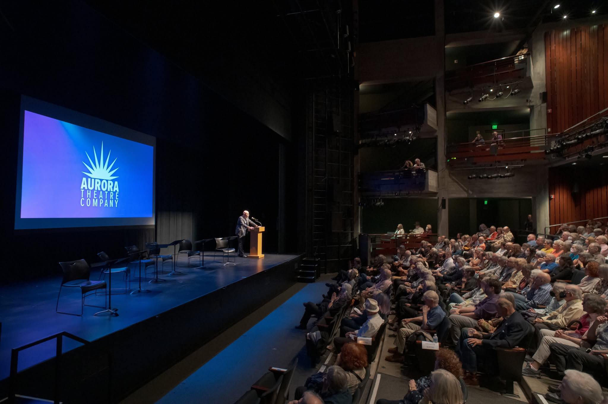 The image shows a theater auditorium with an audience seated facing a stage. On the stage, there is a large screen displaying the logo of Aurora Theatre Company, with a man speaking at a podium.