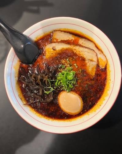 Bowl of spicy ramen with slices of pork, seaweed, green onions, an egg, and red broth, with a black spoon resting inside.