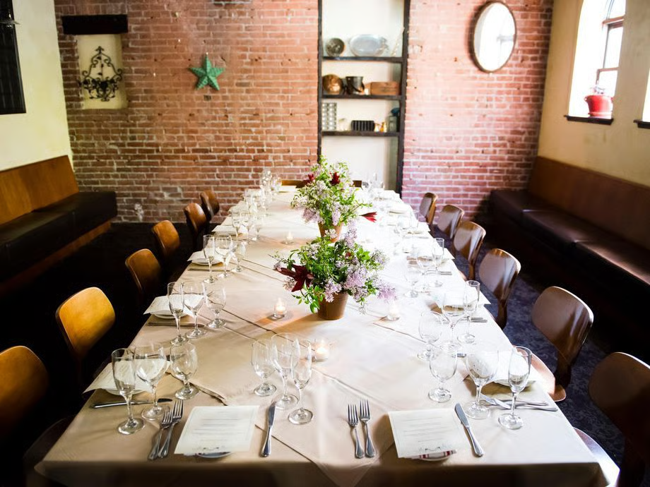 A long dining table set for a meal with white tablecloth, arranged with wine glasses, silverware, napkins, and a floral centerpiece, in a restaurant with brick walls, wooden chairs, and windows.