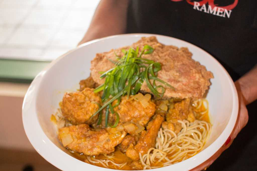 A white bowl filled with ramen noodles topped with fried chicken, a slice of fried pork cutlet, and garnished with sliced green onions.
