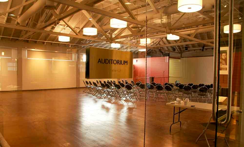 Empty auditorium with chairs arranged in rows, large screens on the wall, and a marked sign reading "AUDITORIUM."