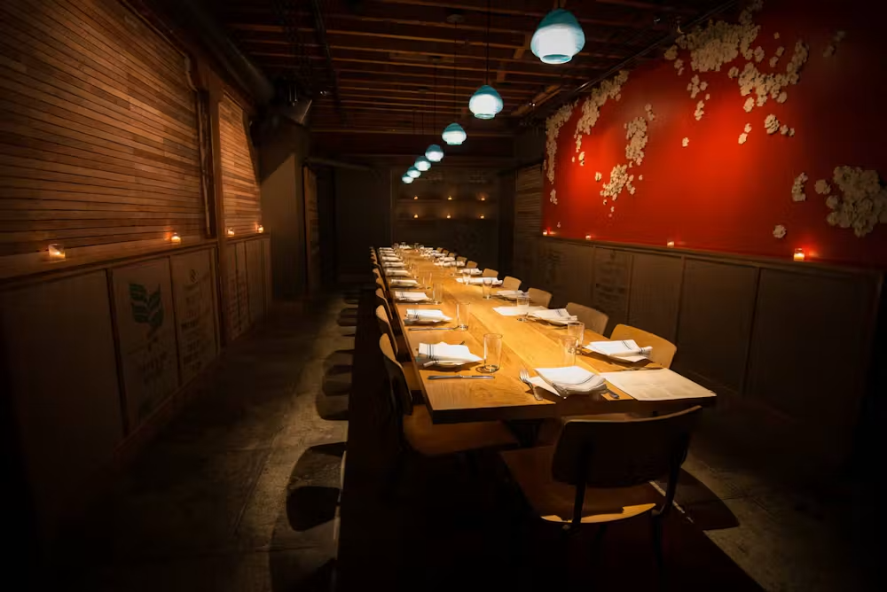 A dimly lit restaurant with a long wooden table set for a meal, with plates, glasses, and napkins arranged neatly. Candles line the walls and a vine pattern decorates a red wall on the right.