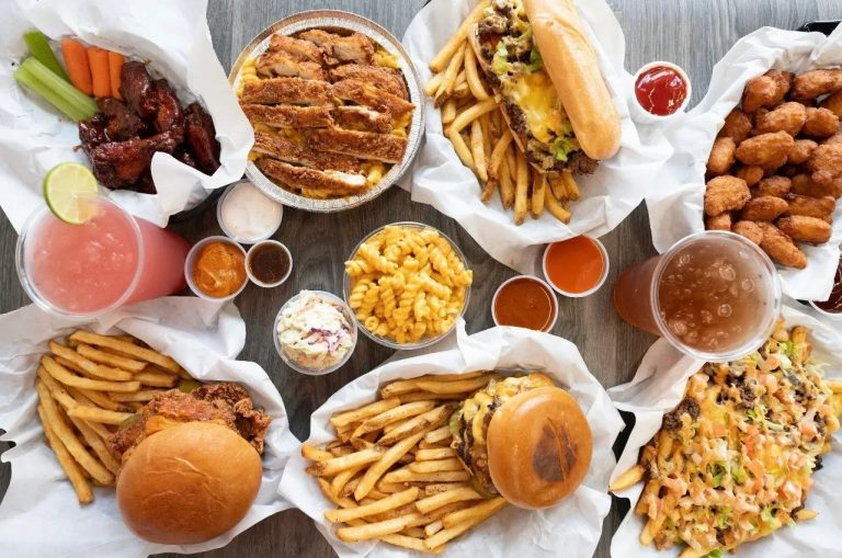 A table with various fast food dishes including fried chicken, French fries, cheeseburgers, loaded fries, pizza, chicken wings, and drinks, with ketchup and sauces.