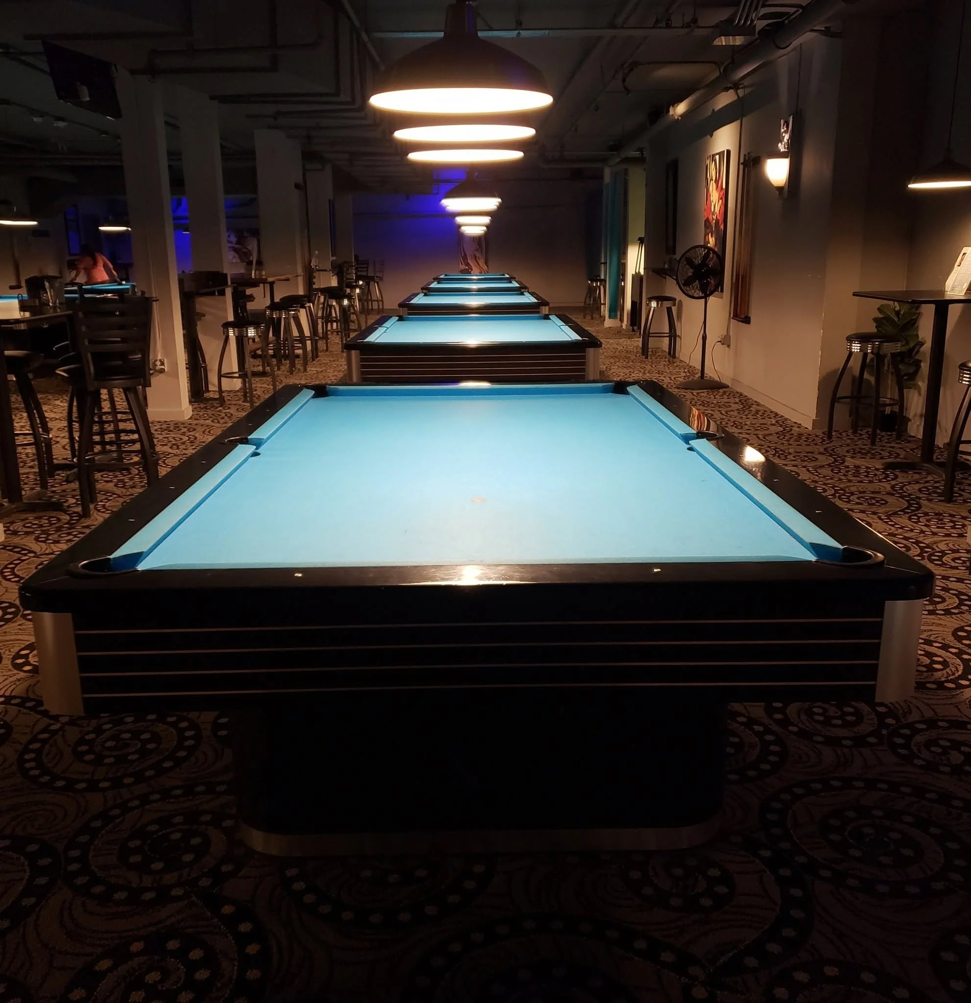 Interior view of a dimly lit pool hall with multiple pool tables illuminated by hanging pendant lights. There are stools and tables along the sides, and a person can be seen in the background.