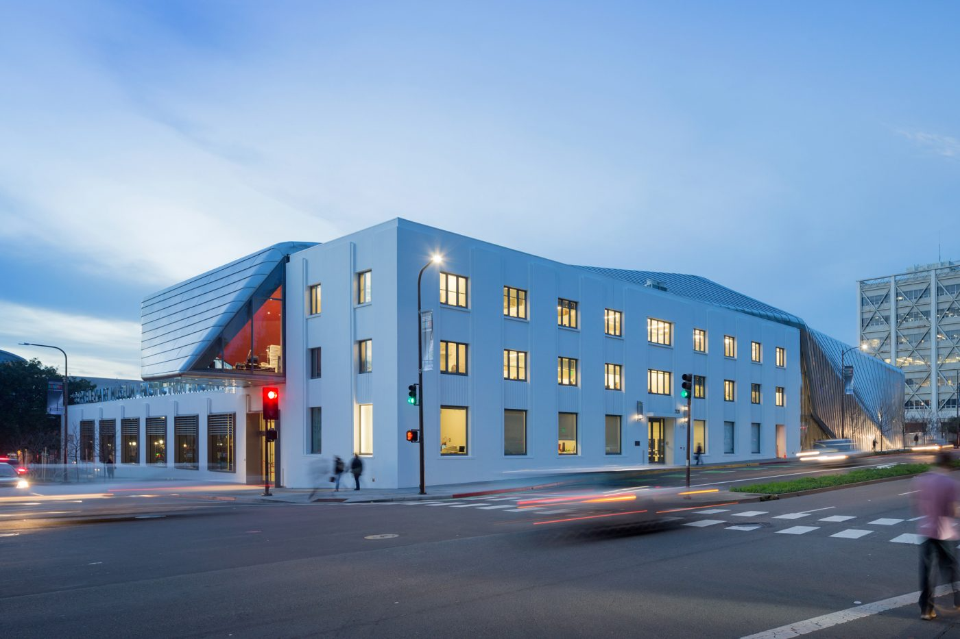 Modern white multi-story building with large windows and metal accents, illuminated at dusk, with traffic lights and pedestrians at an intersection.