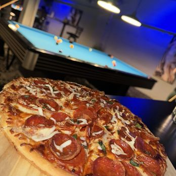 A pepperoni and sausage pizza on a wooden serving board with a pool table in the background.