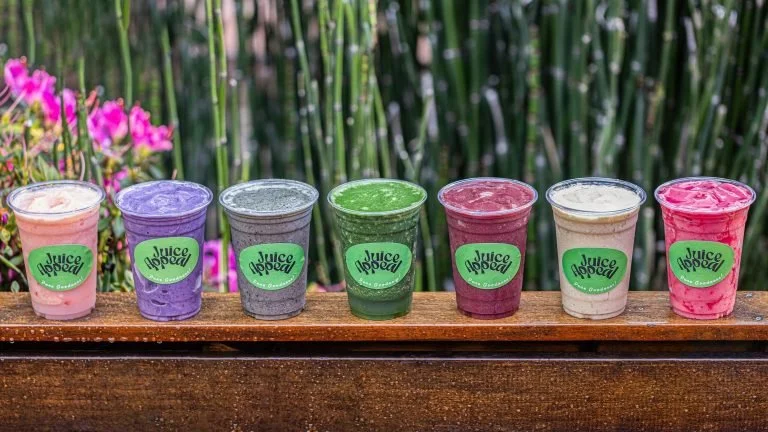 Seven colorful smoothies in plastic cups with green Juice Head labels on a wooden ledge with pink flowers and a green bamboo background.